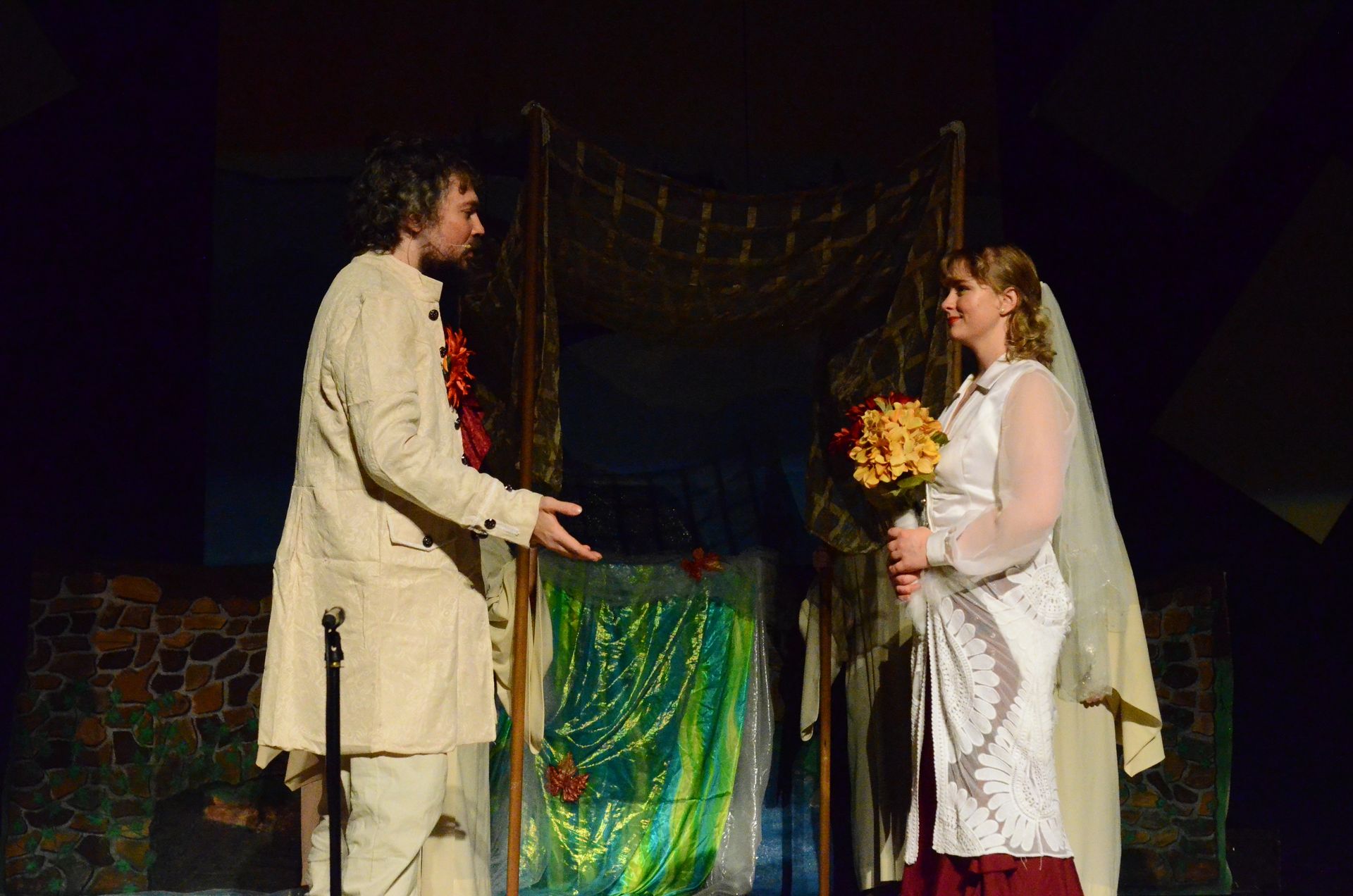 Two performers in cream-colored costume robes stand facing each other on stage, with one holding a bouquet of yellow flowers and the other holding red flowers, in front of a wooden structure decorated with green fabric and gold garland.