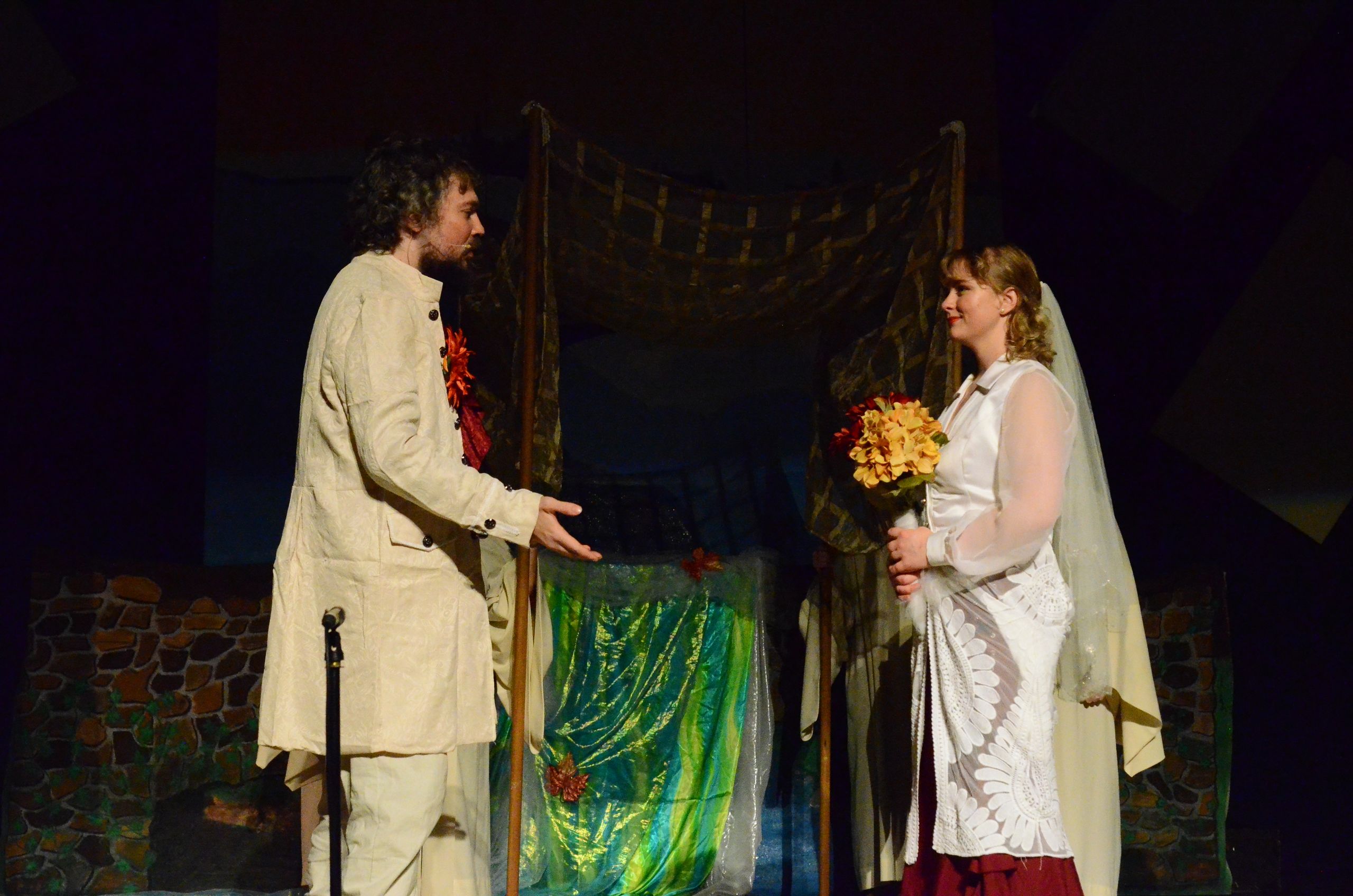 Two performers in cream-colored costume robes stand facing each other on stage, with one holding a bouquet of yellow flowers and the other holding red flowers, in front of a wooden structure decorated with green fabric and gold garland.