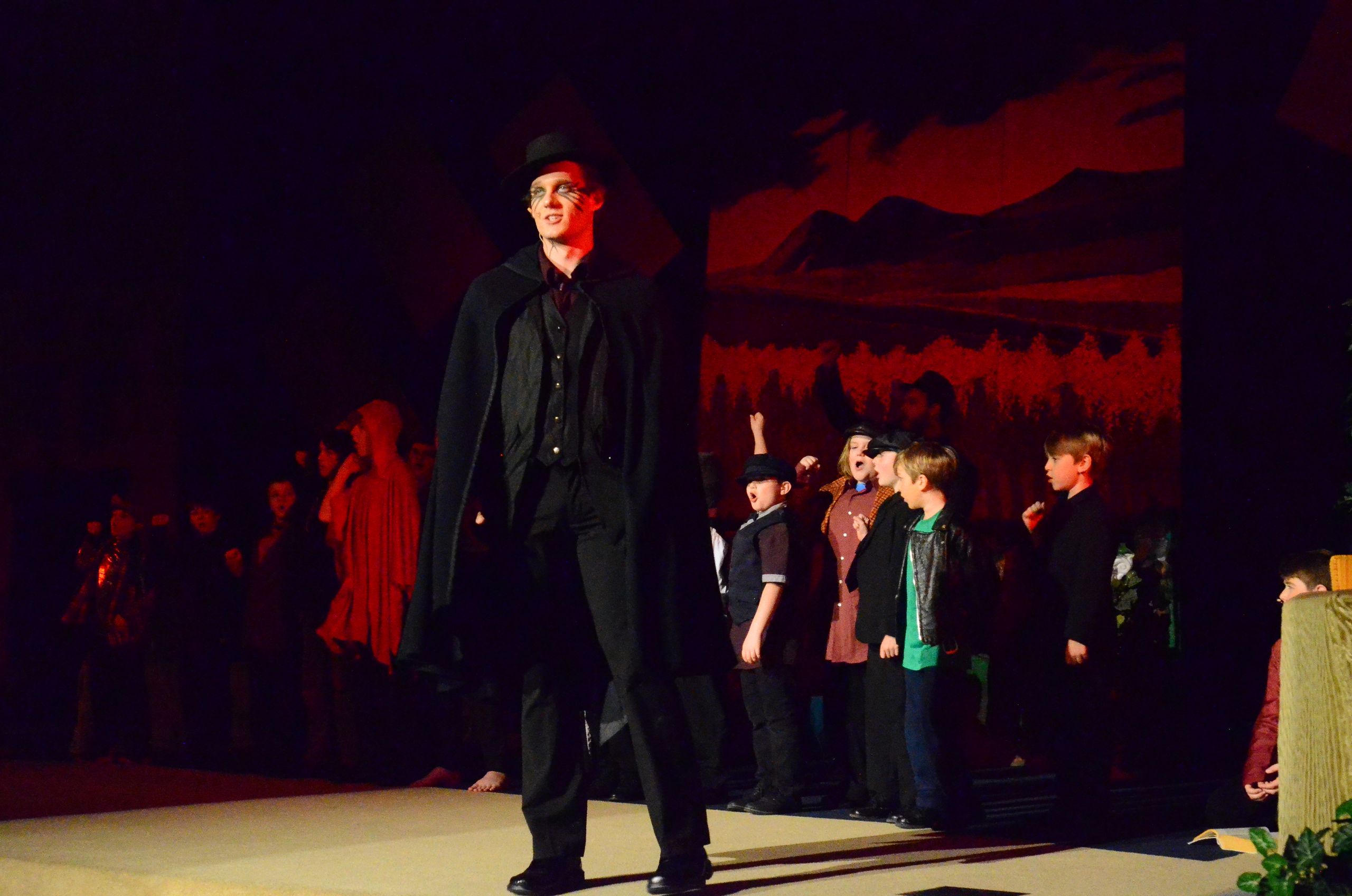 A tall performer in a black coat and hat stands center stage while smaller performers in various costumes surround him against a dramatic red-lit backdrop depicting a crowd scene.