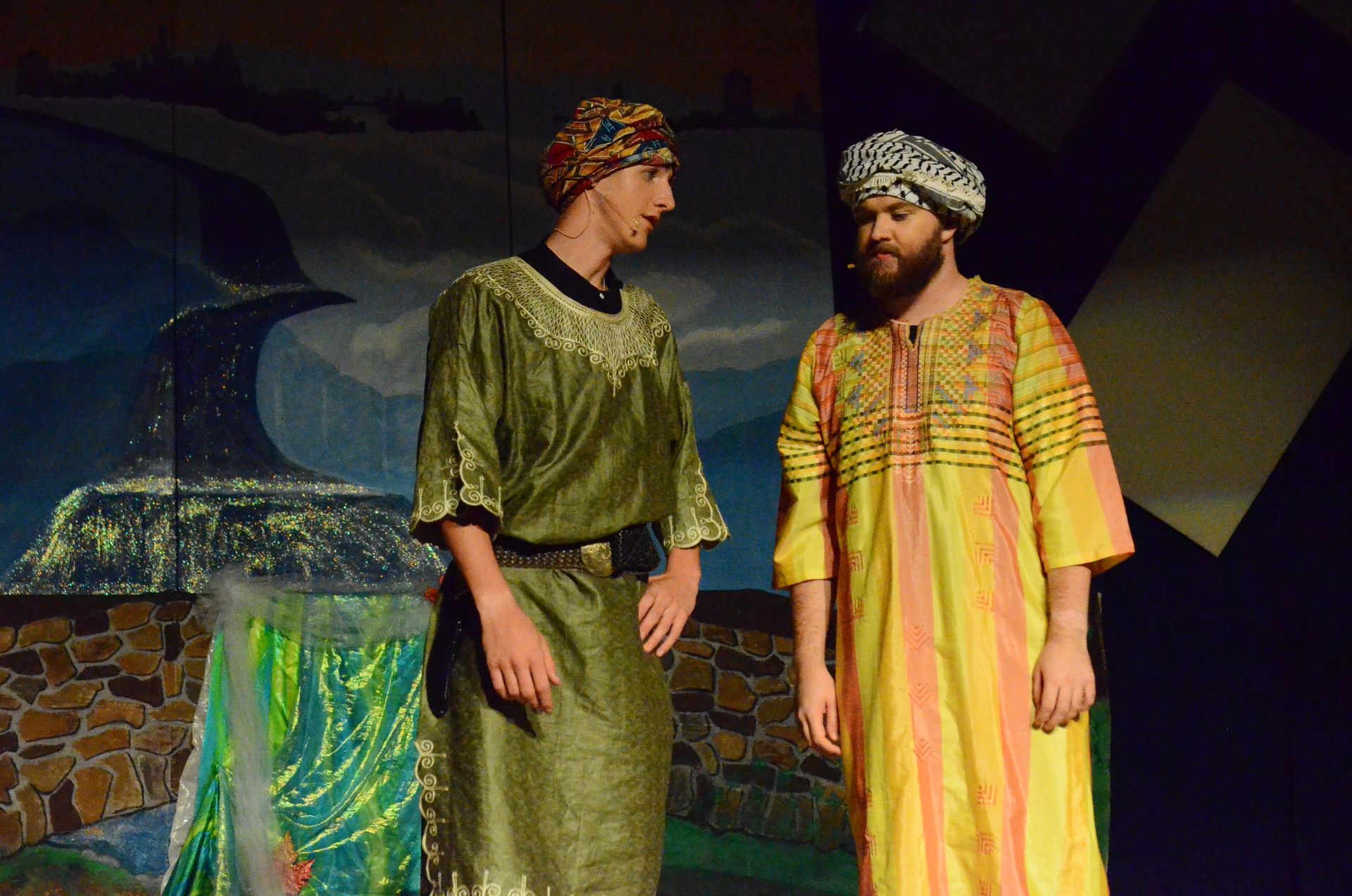 Two performers in colorful tunics and patterned headscarves stand on a stage set with stone walls and a painted backdrop depicting a coastal fortress, engaged in conversation.