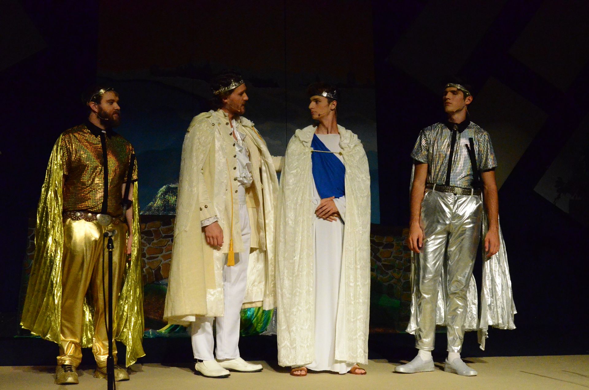 Four performers stand on stage wearing elaborate metallic and flowing robes with decorative capes, posed against a dark backdrop with scenic elements suggesting an ancient setting.