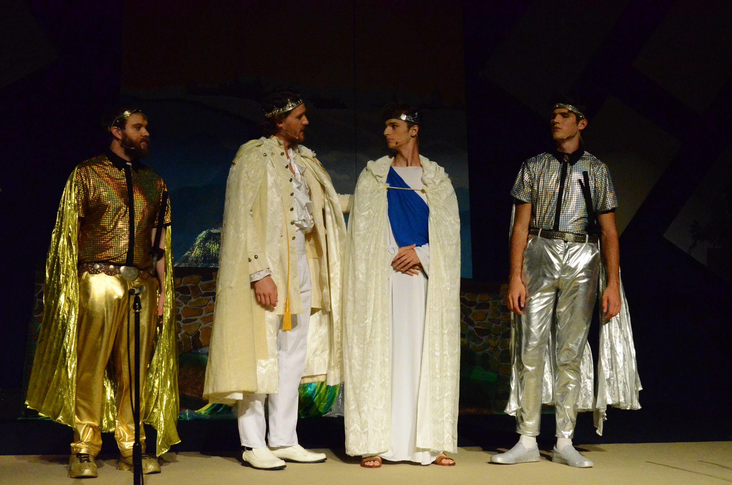 Four performers stand on stage wearing elaborate metallic and flowing robes with decorative capes, posed against a dark backdrop with scenic elements suggesting an ancient setting.