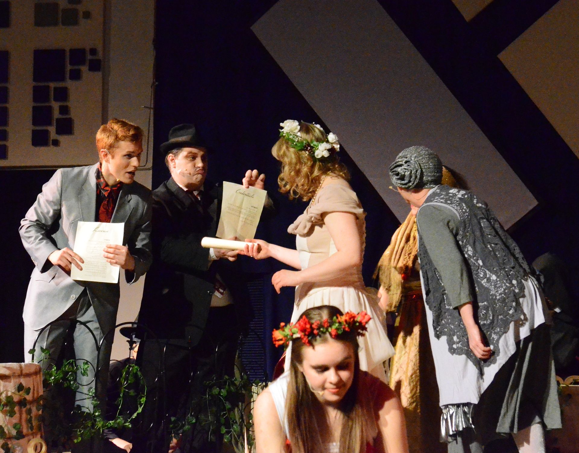 Five performers in period costumes stand on stage around a seated performer wearing a flower crown, with the standing performers exchanging scrolls and documents while geometric set pieces frame the scene behind them.