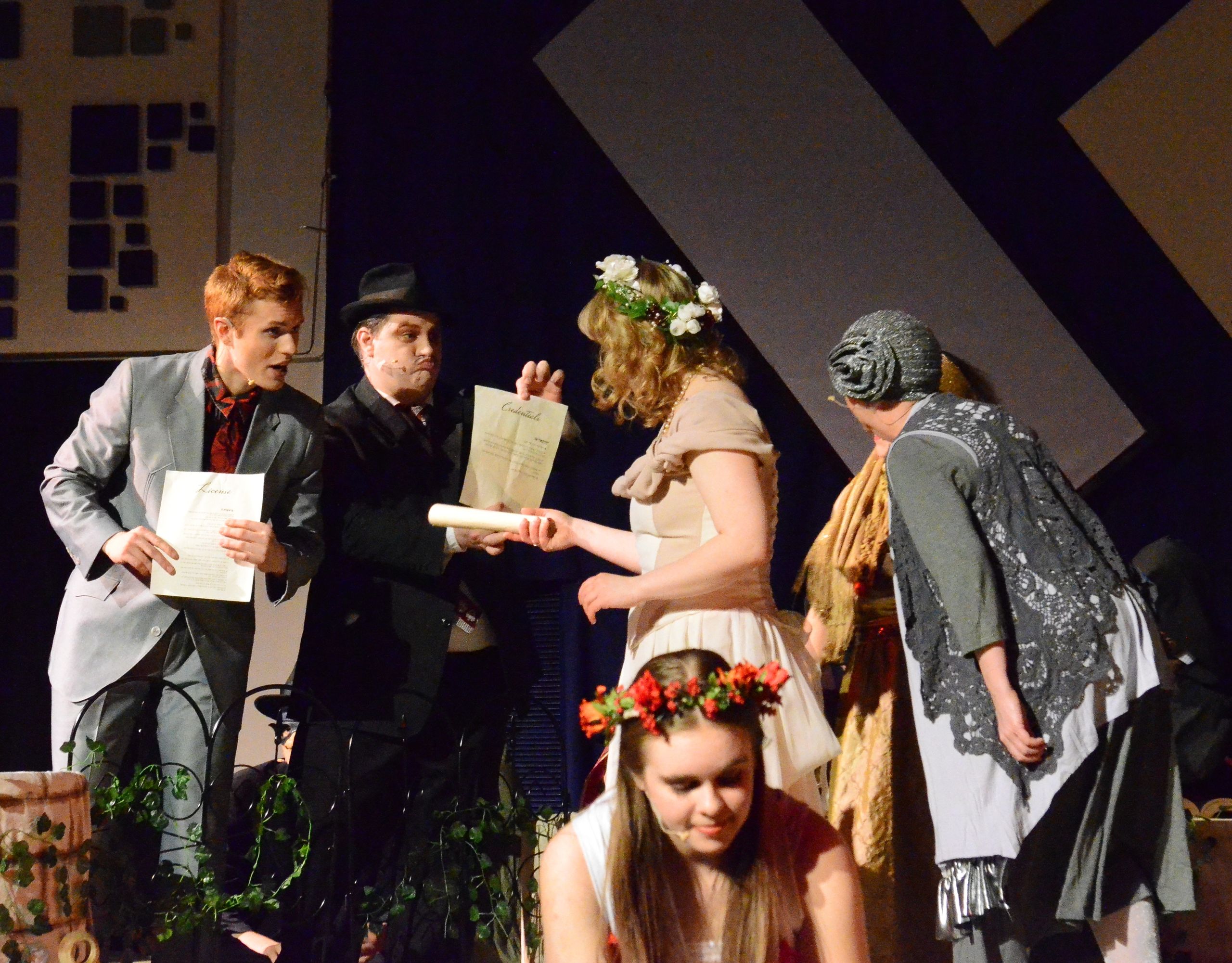 Five performers in period costumes stand on stage around a seated performer wearing a flower crown, with the standing performers exchanging scrolls and documents while geometric set pieces frame the scene behind them.