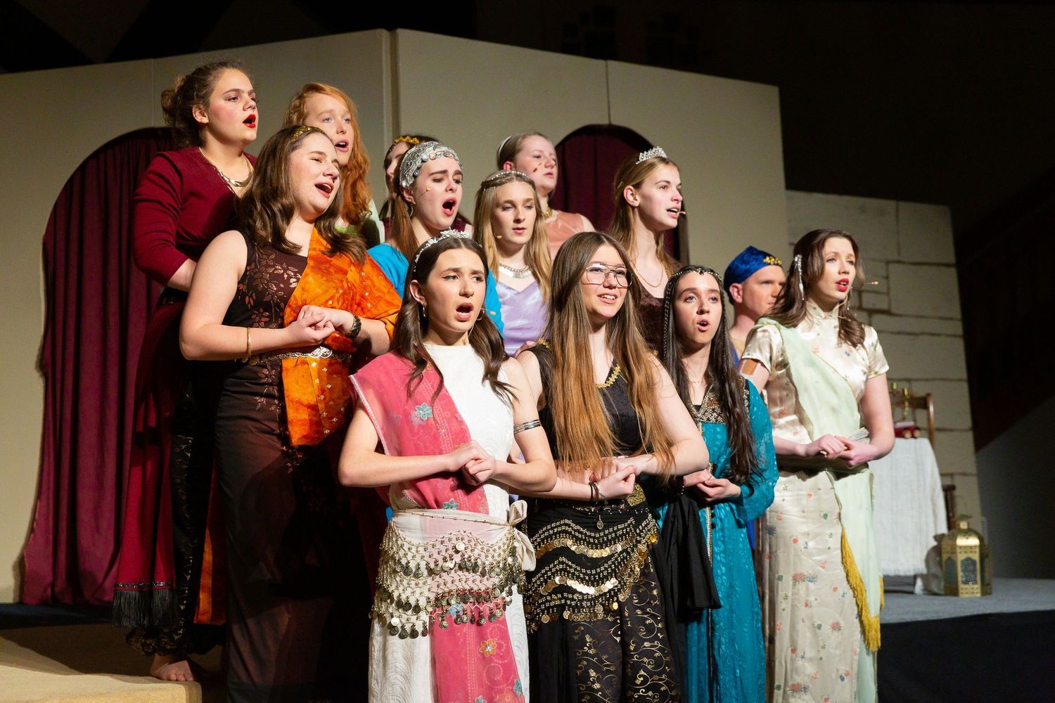 A diverse cast of performers in ornate costumes featuring beaded embellishments, colorful fabrics, and decorative headpieces stands together on stage singing, with a neutral backdrop and stage lighting behind them.