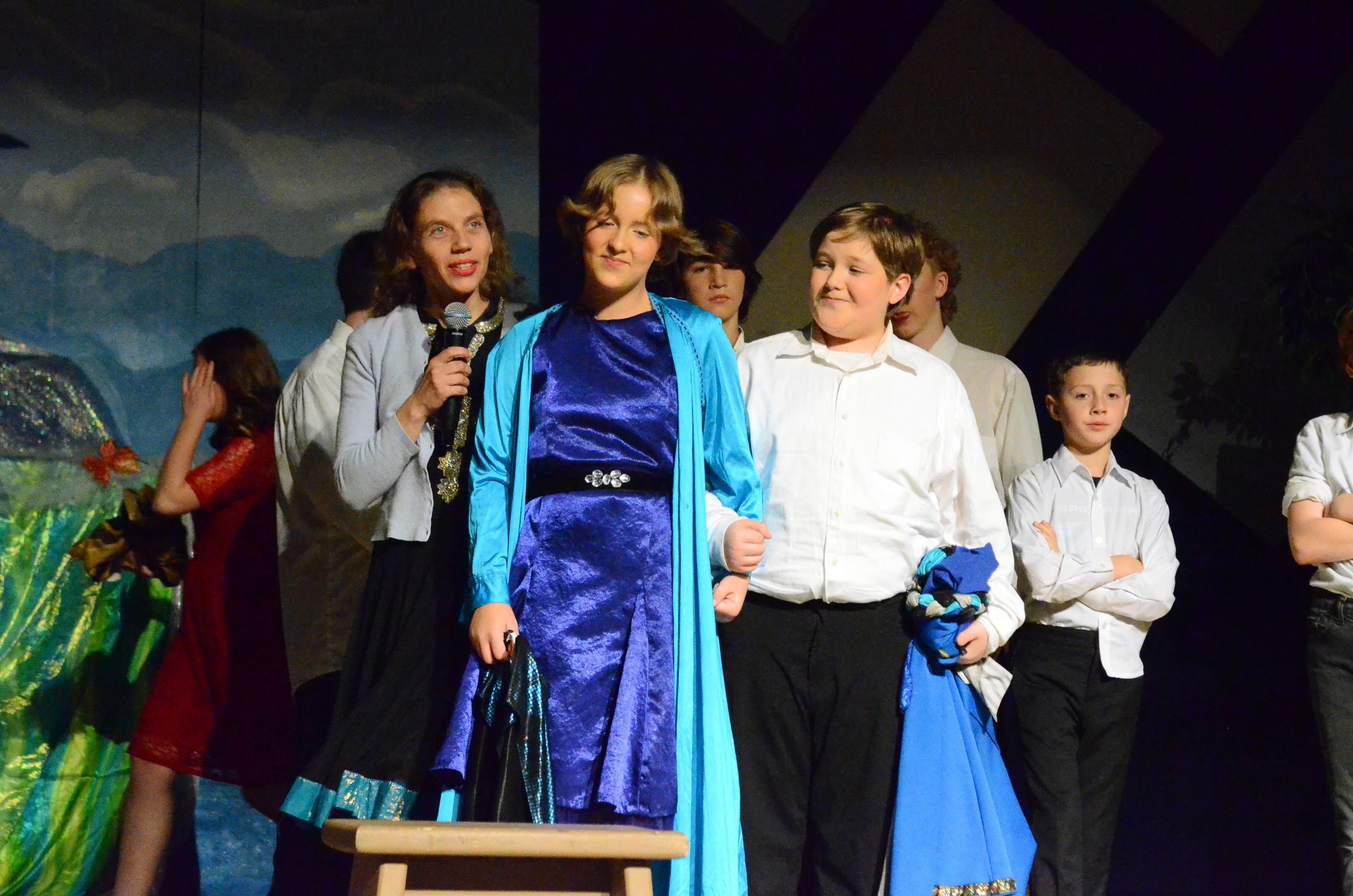 A group of performers stands on stage in costume, with one wearing a striking blue velvet dress and matching shawl standing center stage holding a microphone, surrounded by cast members in period clothing against a painted backdrop.