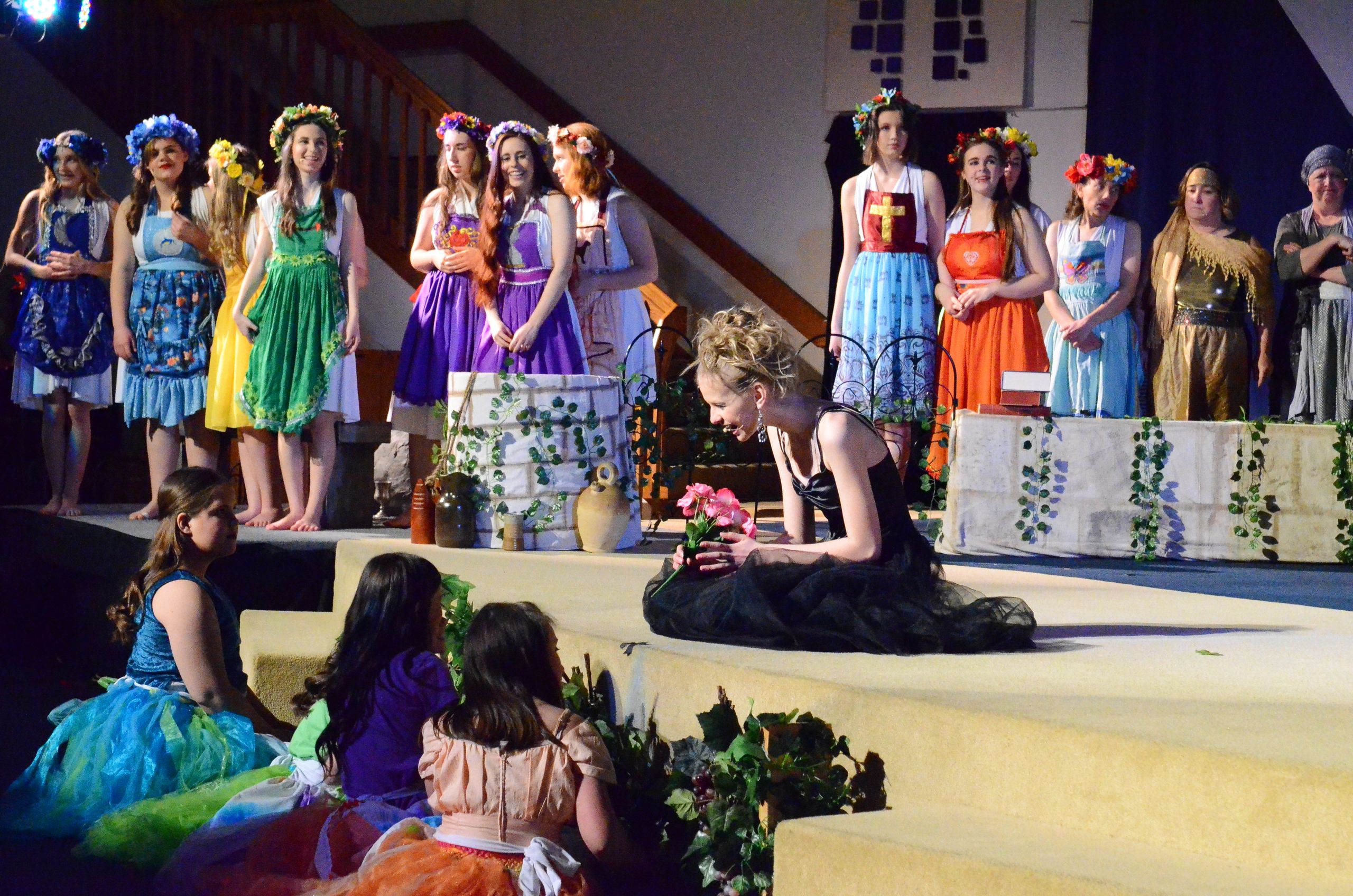 A cast of performers in colorful floral-themed costumes and flower crowns stands on an elevated stage while three performers in the foreground sit on the lower stage level, with decorative potted plants and ivy-draped set pieces visible throughout the scene.