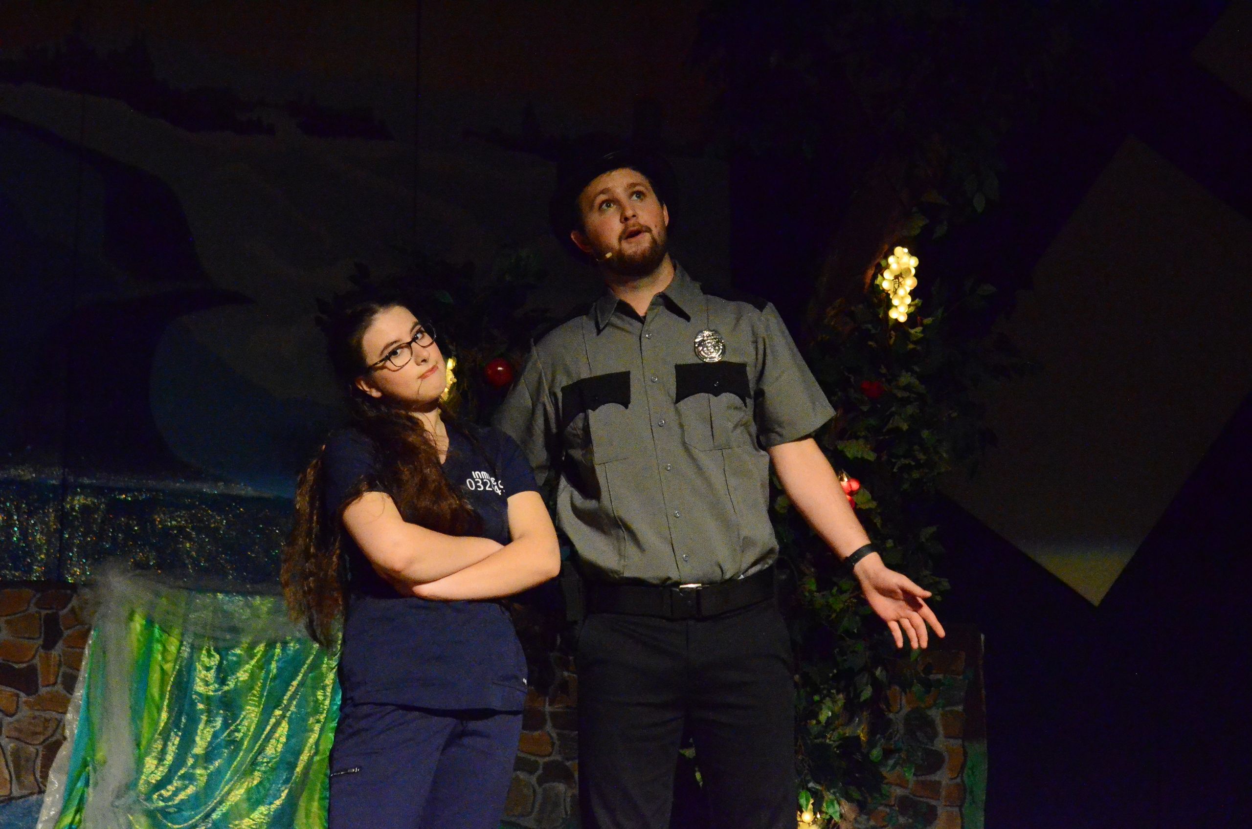 Two performers stand on a dimly lit stage decorated with green fabric and ivy, with one wearing a ranger-style uniform and the other in a navy blue shirt, both gazing upward toward a glowing light source above them.