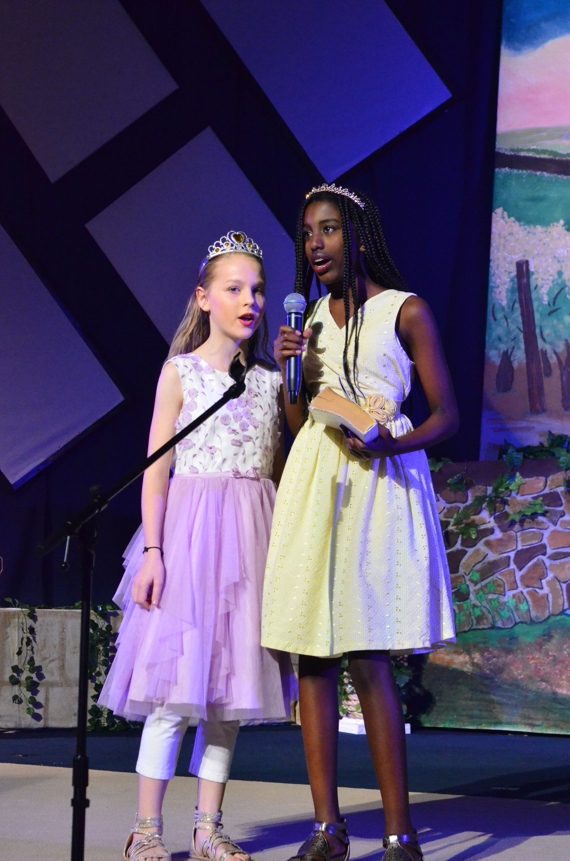 Two performers in elegant formal attire—one wearing a yellow dress and the other in a lavender tulle skirt—stand center stage under purple lighting while singing into microphones, with colorful scenic elements and decorative foliage visible in the background.