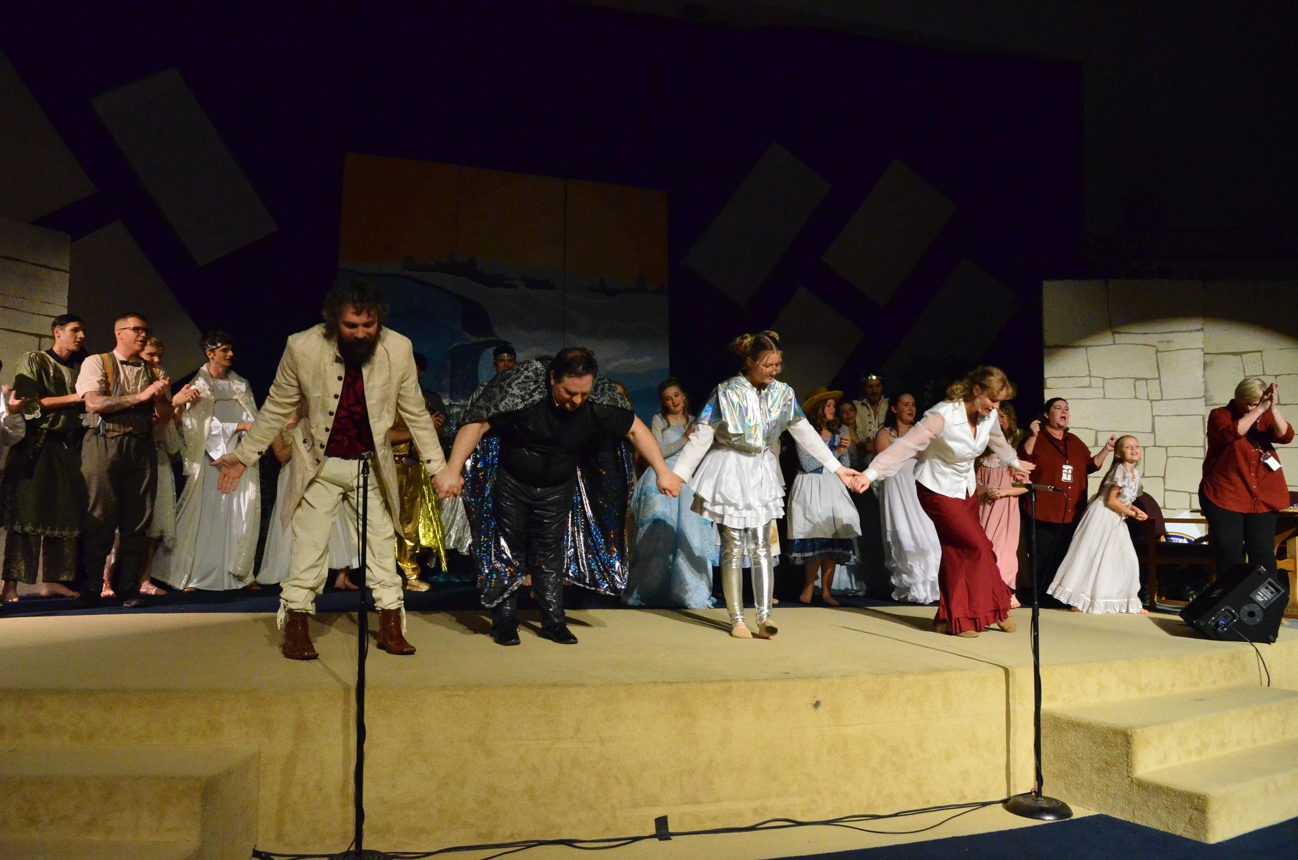 The cast takes a bow on stage in period costumes including earth tones, whites, and reds, with stylized set pieces and a painted backdrop visible behind them.