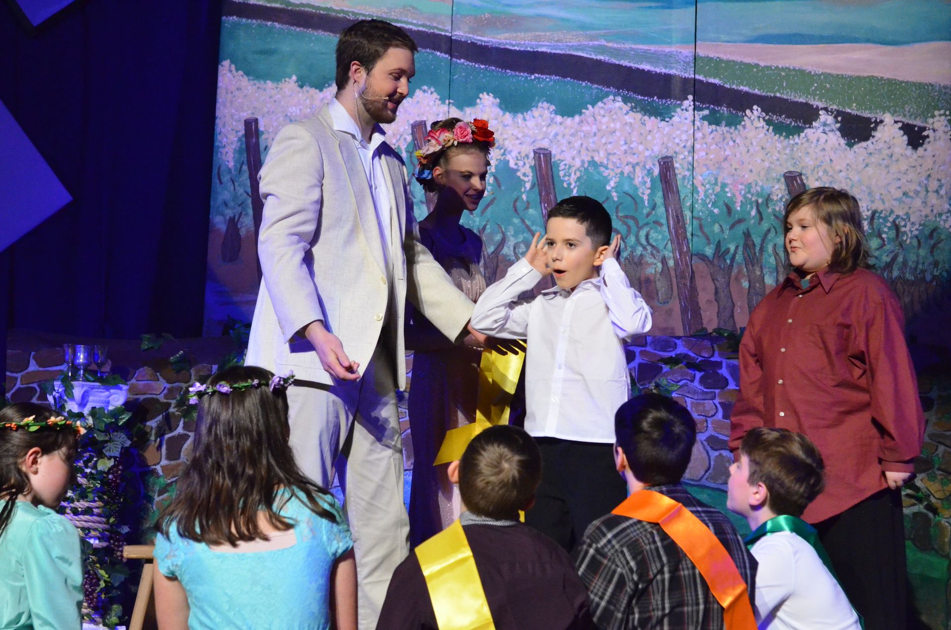 A cast of performers in period costumes interact with young audience members on stage in front of a painted backdrop depicting a flowering field with distant mountains.