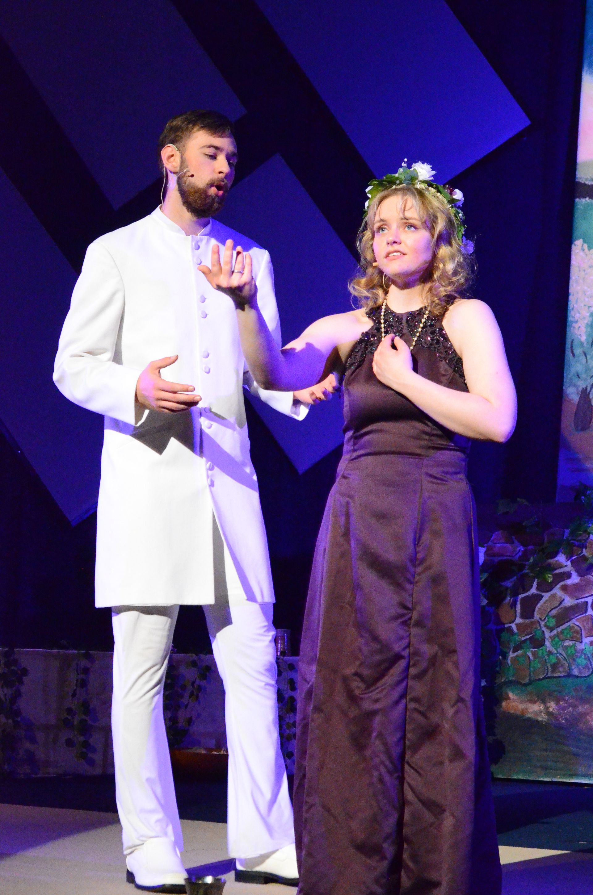 Two performers in formal attire dance together on stage under blue lighting, with one wearing a dark dress with a floral crown and the other in a white suit, against a colorful backdrop.