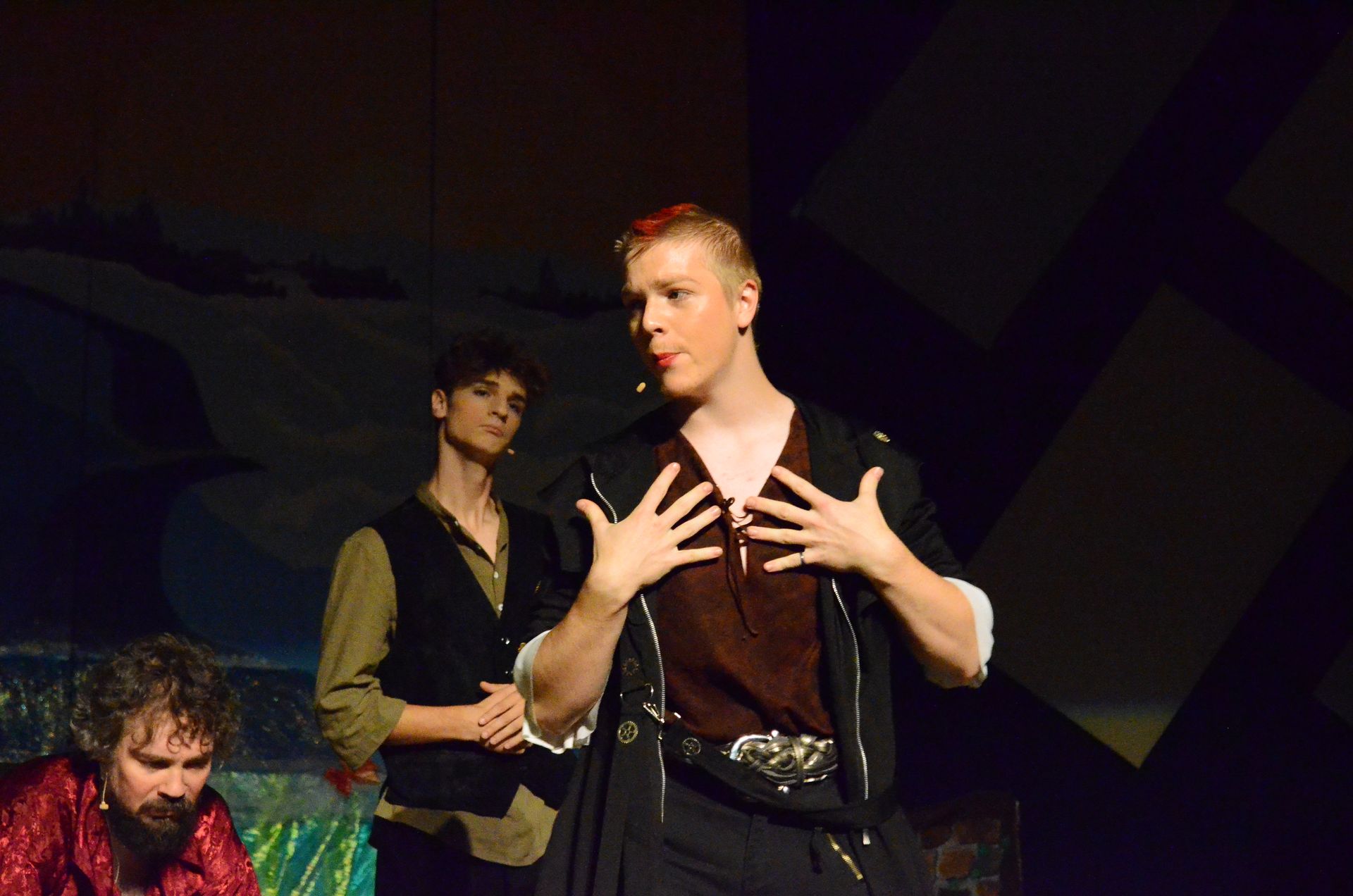 Three performers on stage wear earth-toned medieval-style costumes against a dark backdrop with colorful scenic elements, as the central performer gestures expressively while speaking or singing.