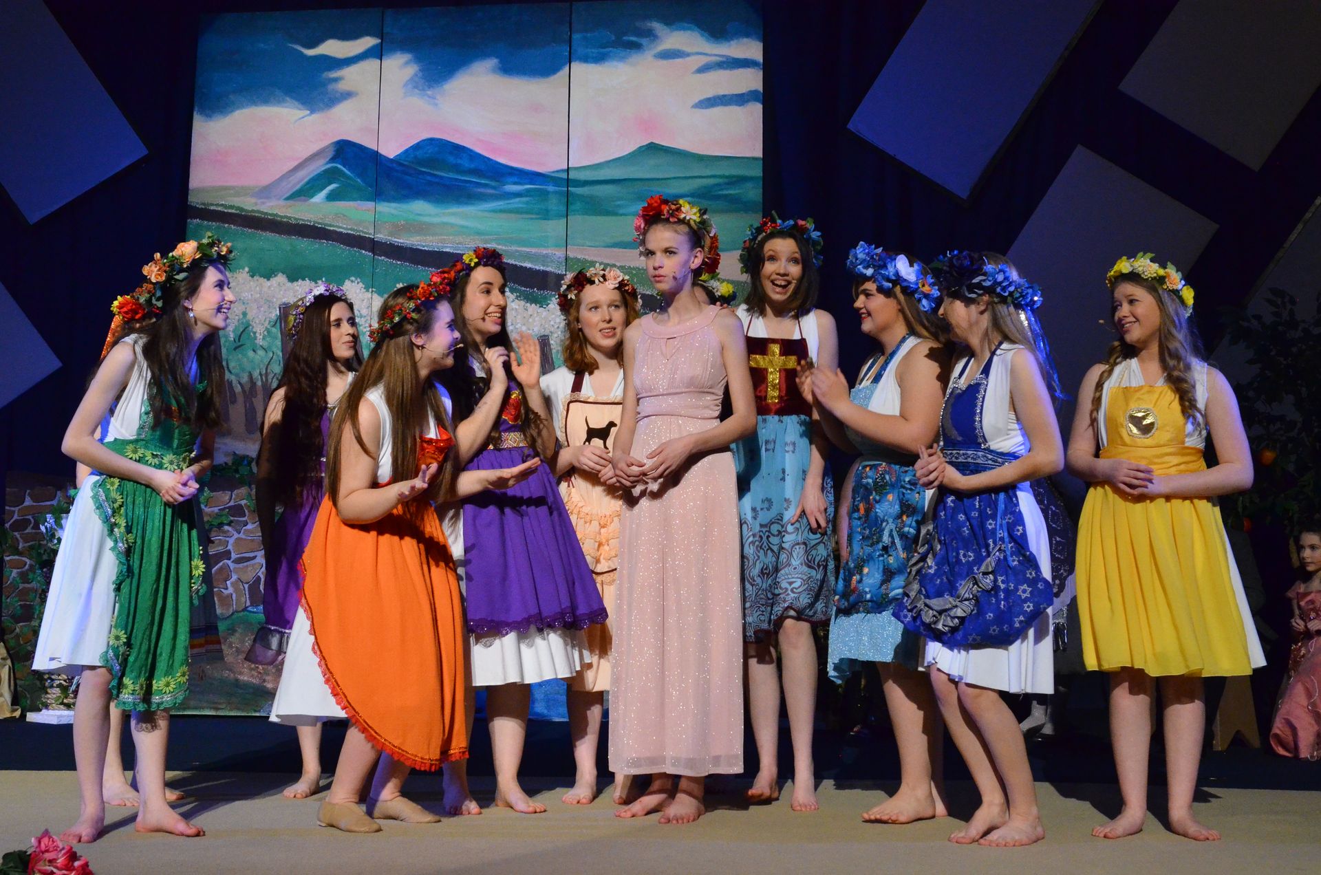 A cast of performers wearing colorful floral crowns and vibrant dresses stands barefoot on stage in front of a painted backdrop depicting mountains and landscape, appearing to be in a scene from a musical production.