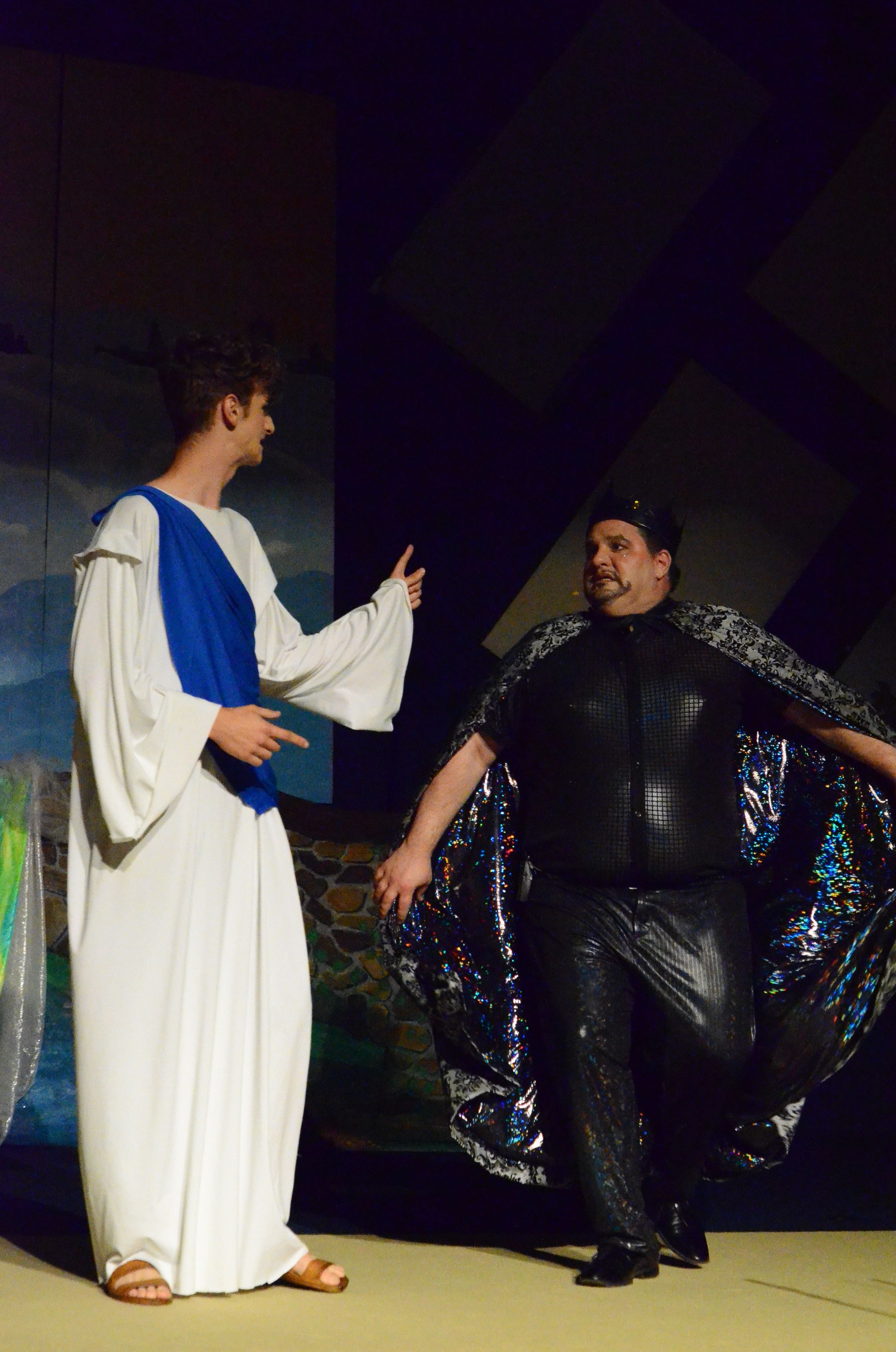 Two performers in sparkly black and blue costumes strike dramatic poses on stage, with one standing upright looking downward and the other lying horizontally in white robes against a blue backdrop.