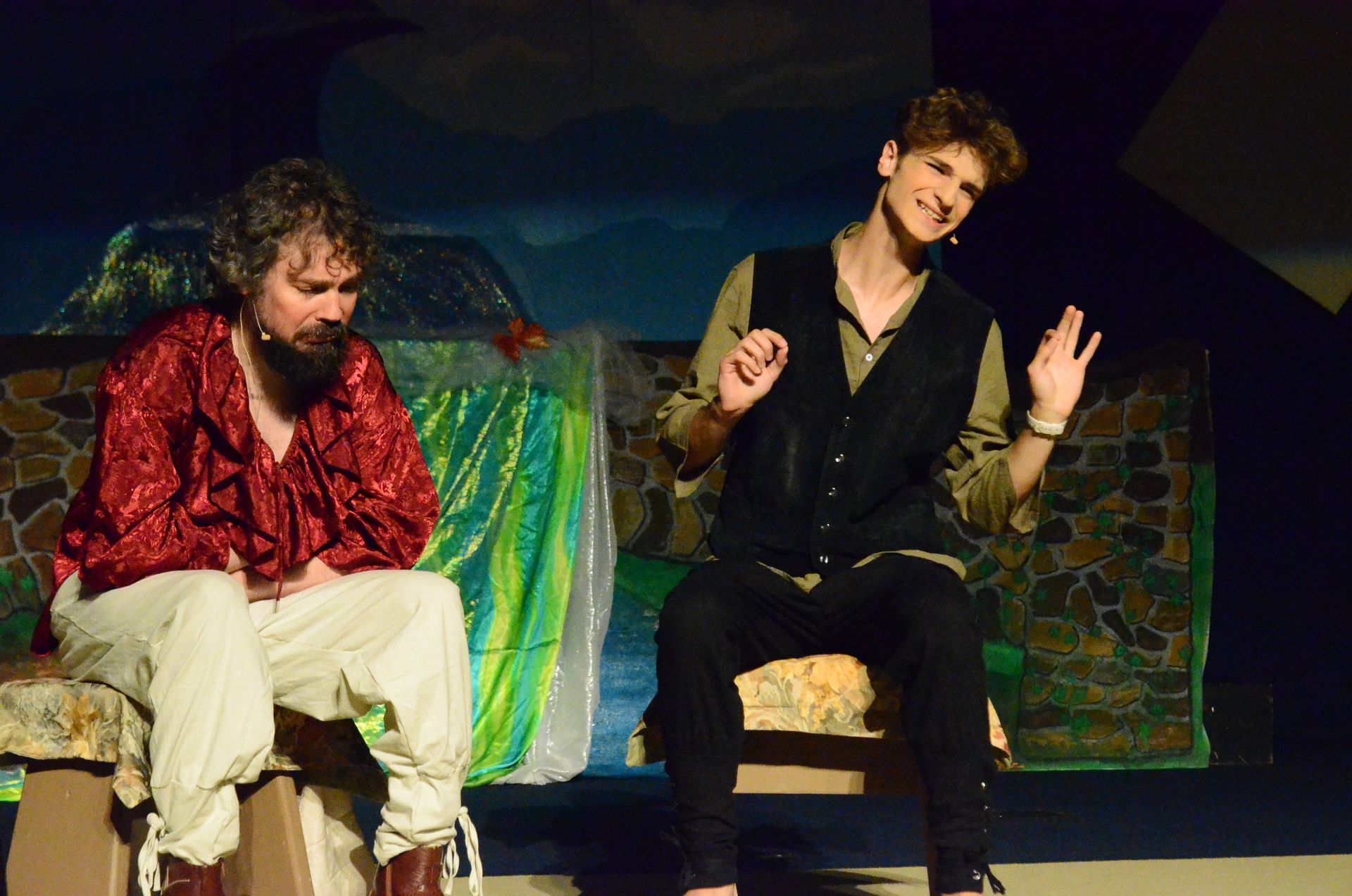 Two performers sit on a wooden bench against a painted backdrop depicting a rustic stone dwelling, with one wearing a red patterned shirt and cream-colored pants while gesturing expressively, and the other in a black vest and olive shirt smiling and gesturing toward their scene partner.