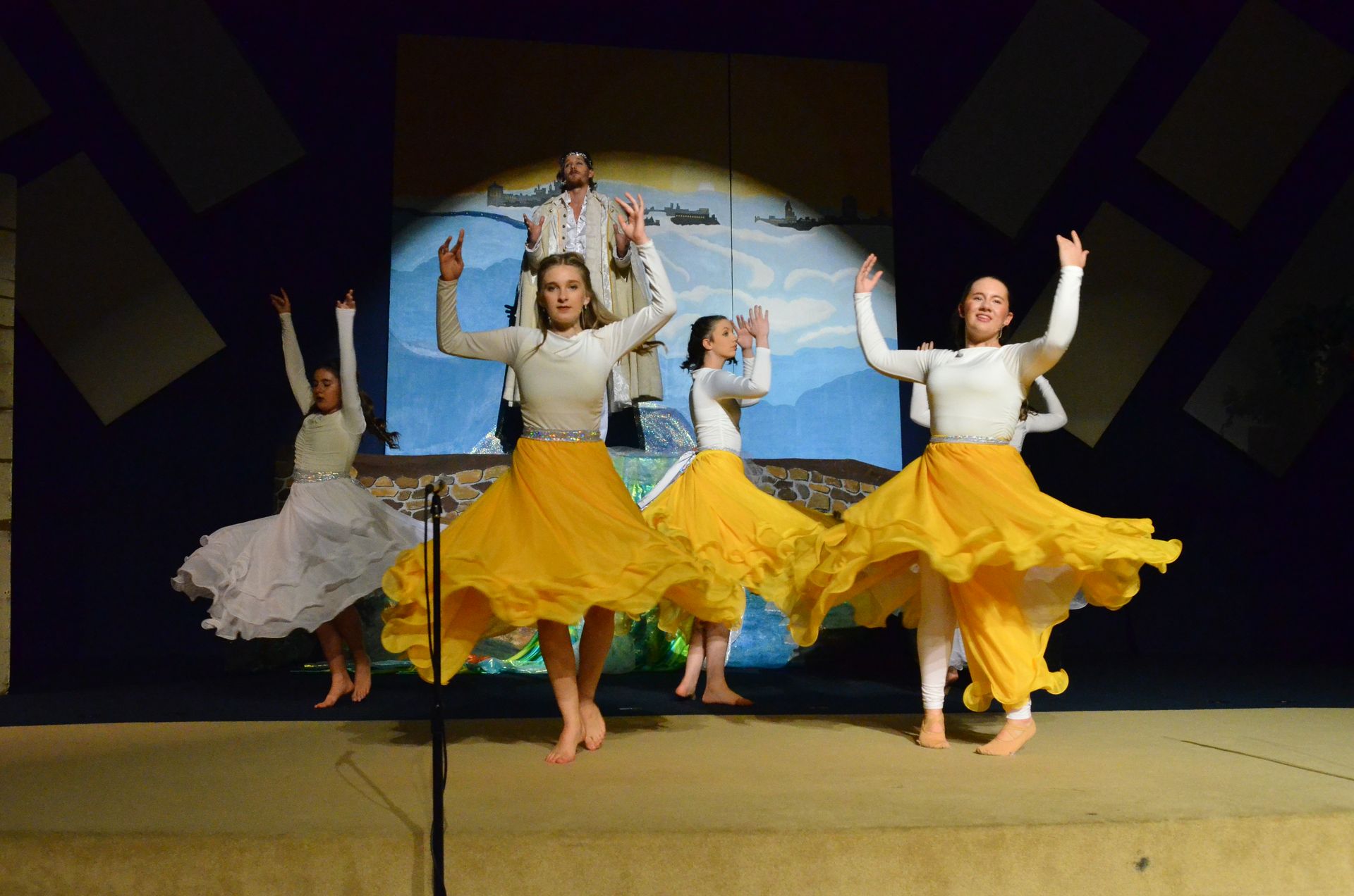 A cast of performers in cream-colored tops and flowing yellow skirts dance with arms raised on stage, with a painted backdrop of sky and landscape visible behind them.