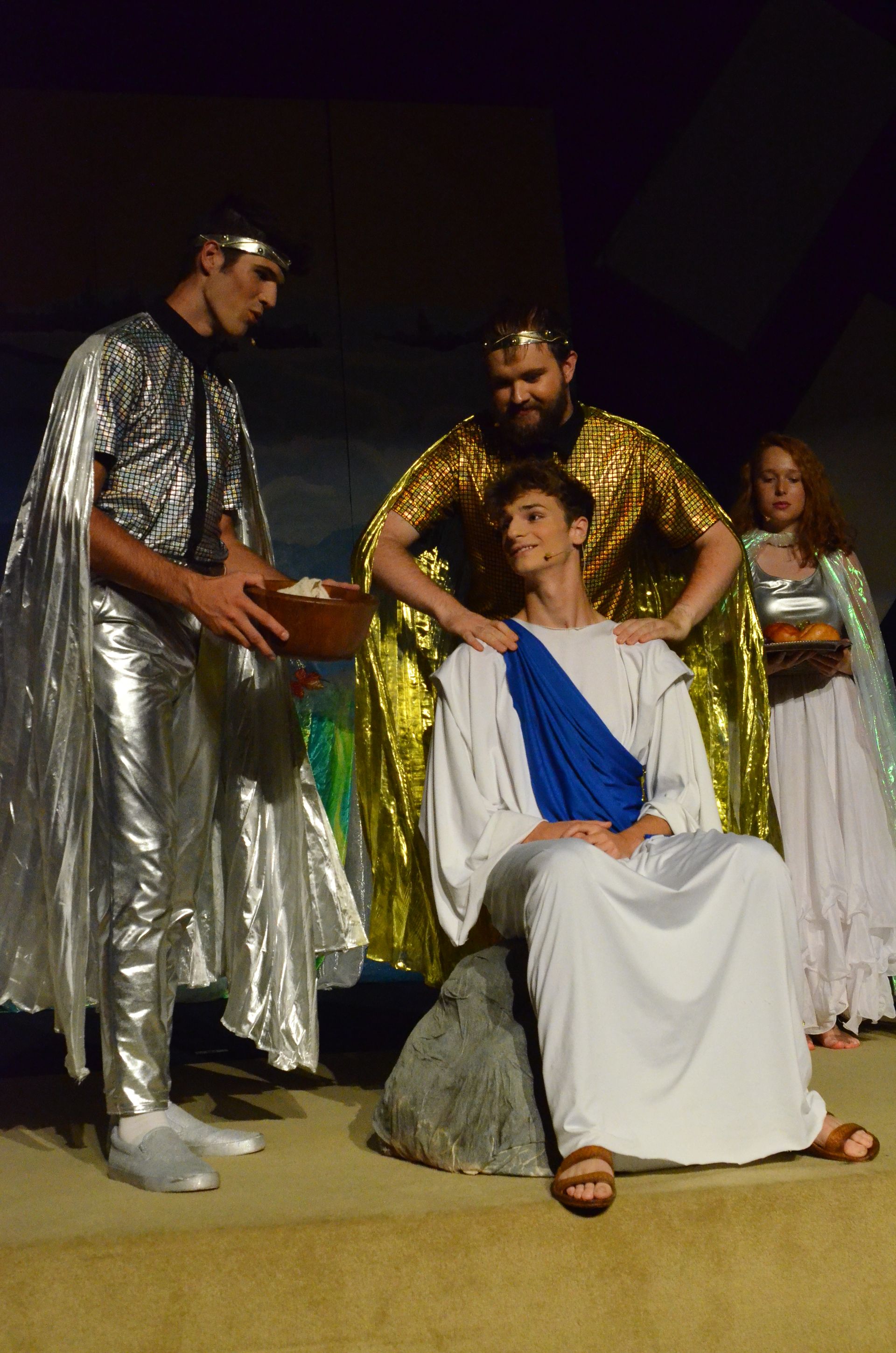 Cast members in metallic gold and silver costumes pose together against a yellow wall, with one performer in white and blue garments standing centrally among the group.