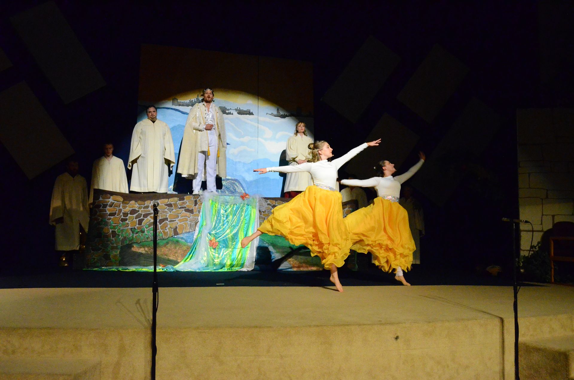 Cast members in white robes stand on a stone platform before a painted backdrop of water and distant ships, while two performers in flowing yellow garments dance expressively in the foreground.