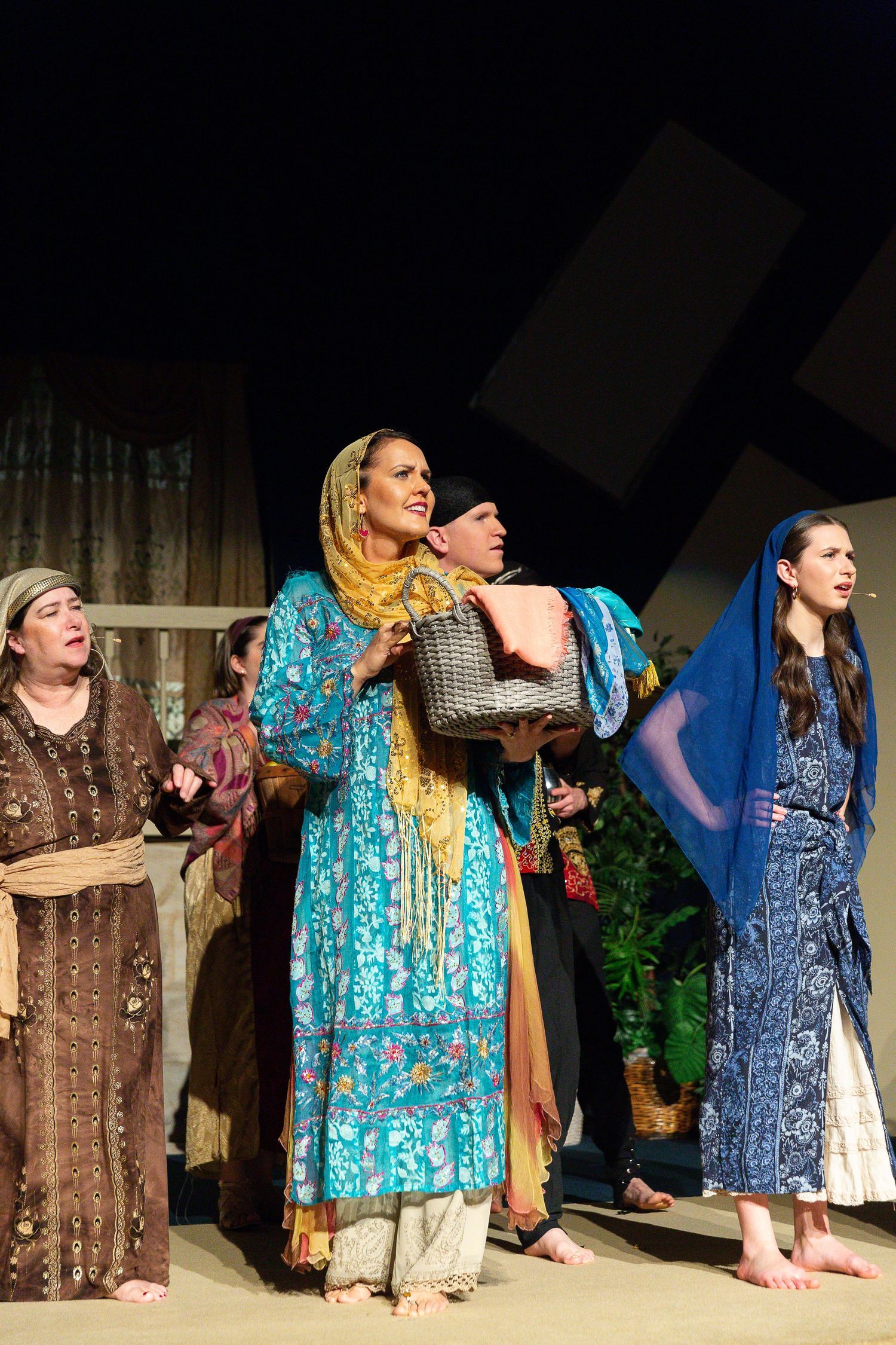 A ensemble of performers in colorful patterned robes and headscarves stands barefoot on stage, gazing upward with expressions of wonder and reverence.