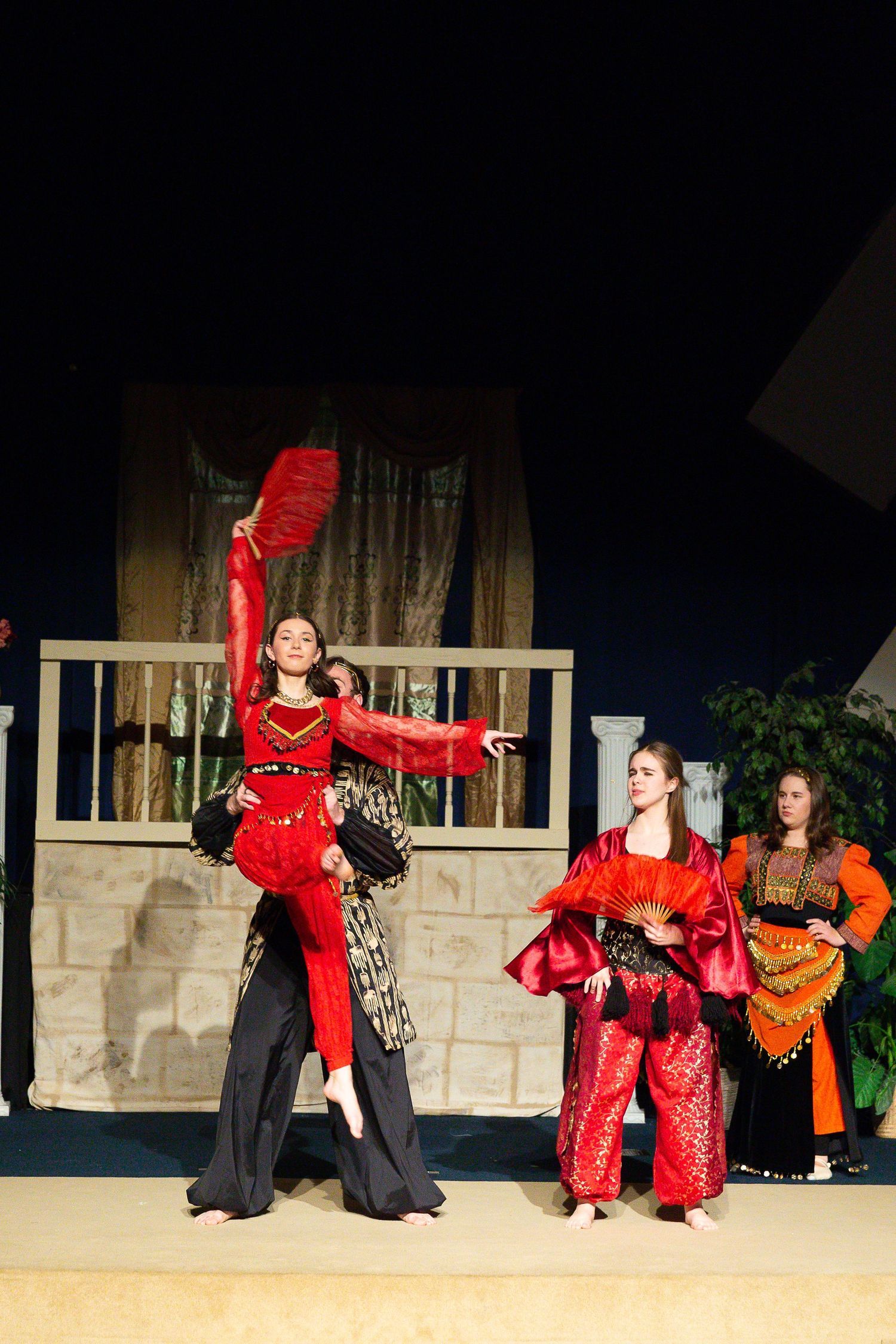 Three performers in elaborate red and gold costumes stand on stage before a wooden structure with a balcony, as one performer gestures dramatically while holding a large red feathered fan.