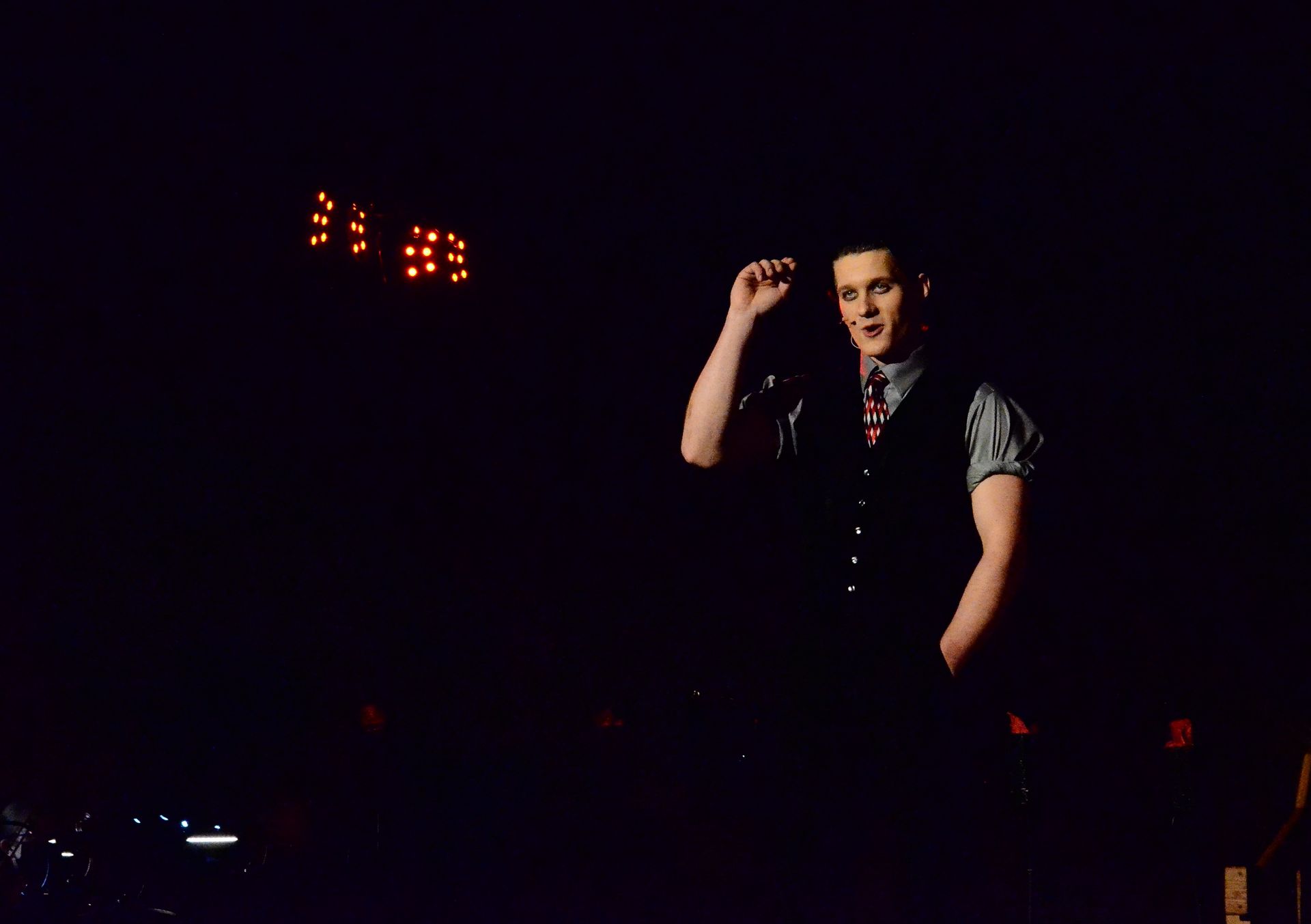 A performer in a gray short-sleeved shirt and black vest with a patterned tie stands spotlit on stage against a dark background, with illuminated marquee lights visible above.