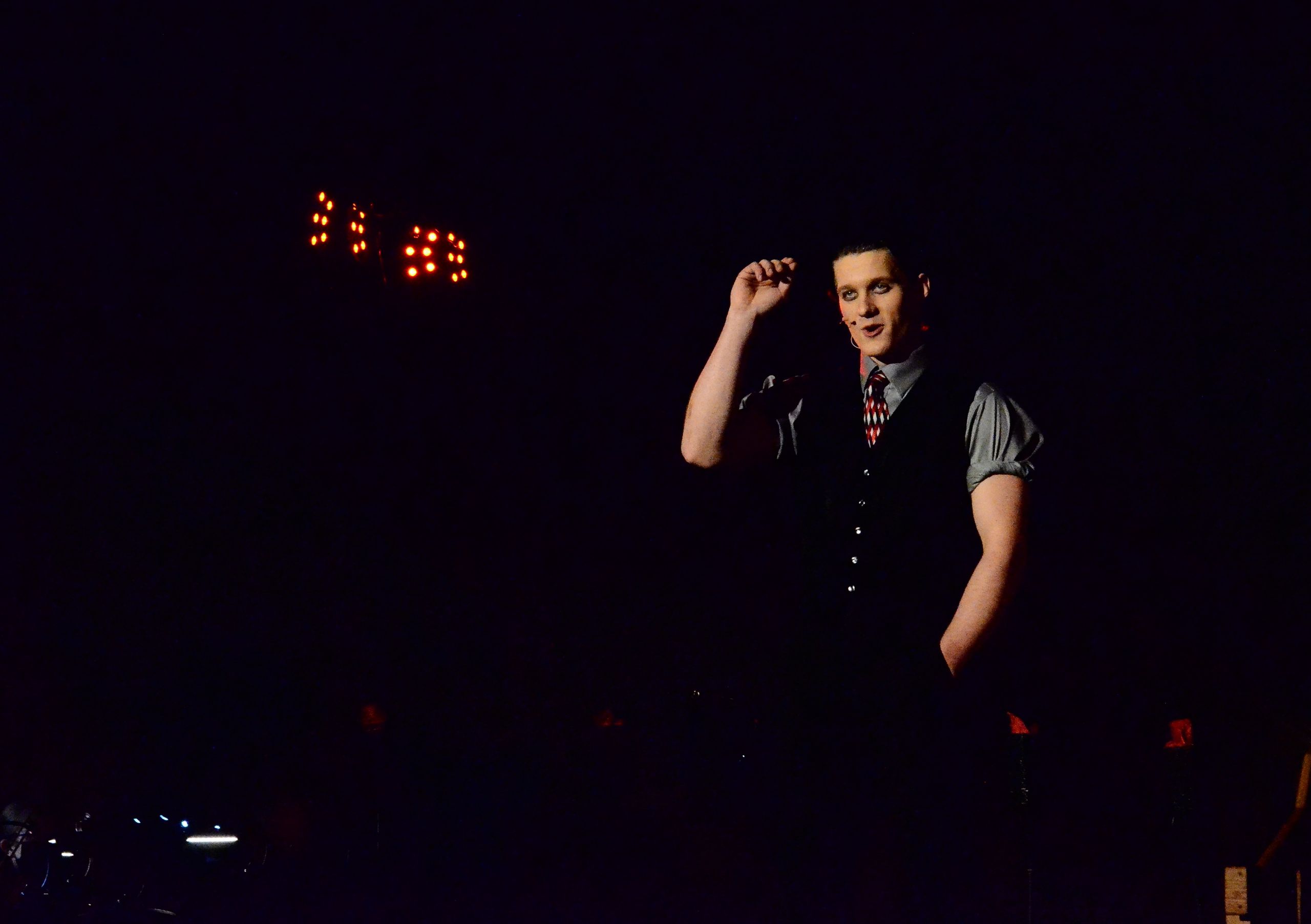 A performer in a gray short-sleeved shirt and black vest with a patterned tie stands spotlit on stage against a dark background, with illuminated marquee lights visible above.