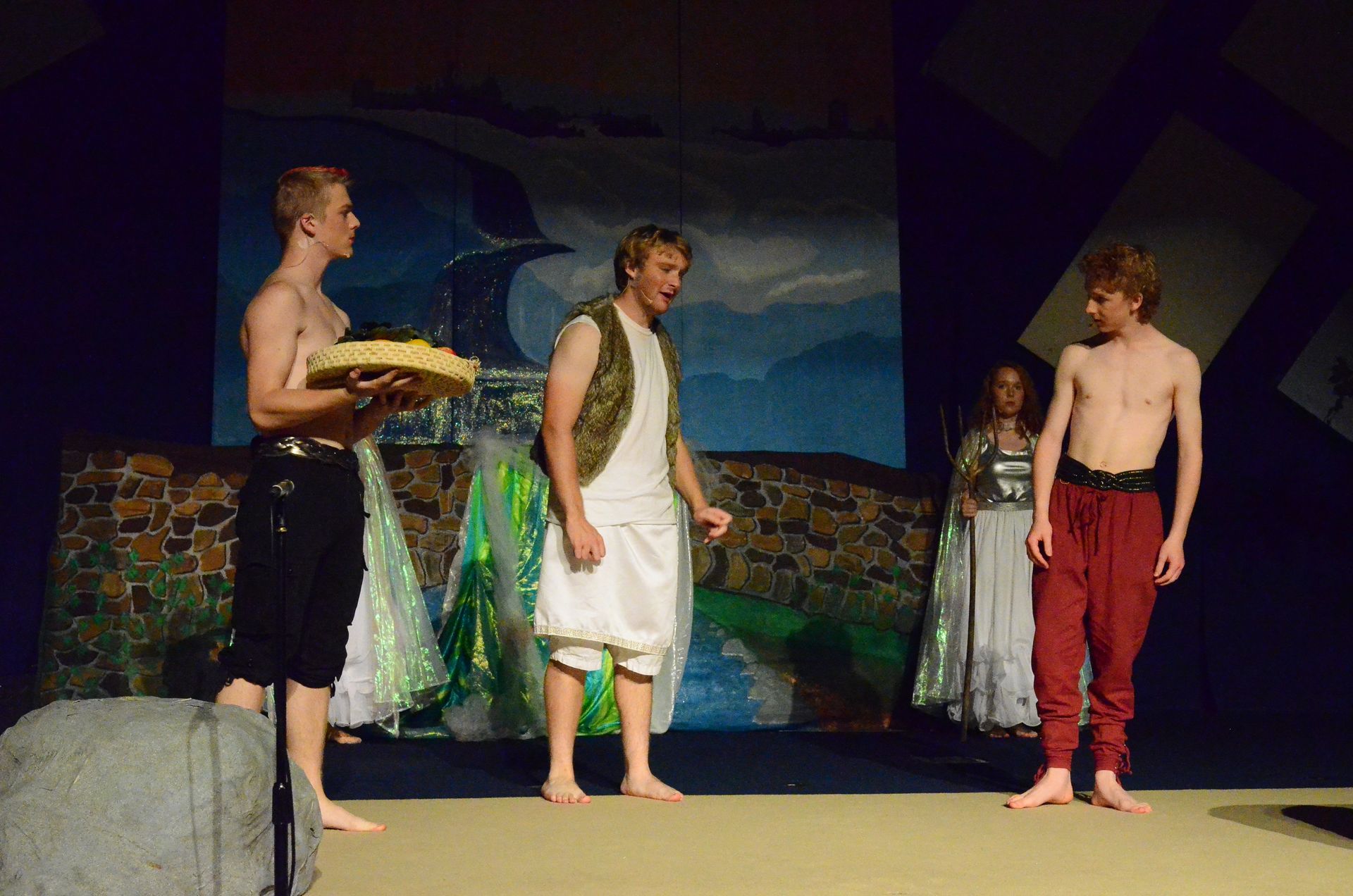 Four performers in classical Greek-style costumes stand on a stage with a painted backdrop depicting a nighttime seascape, with one performer holding a loaf of bread and others wearing flowing green and white garments.