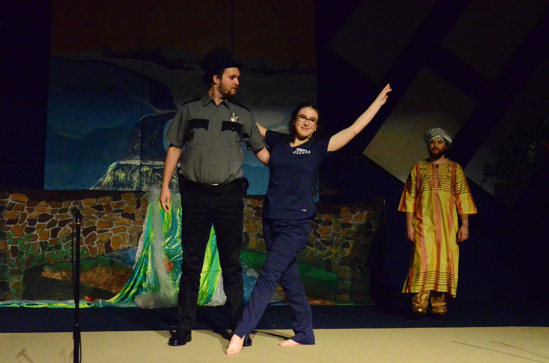 Three performers stand on stage in front a stone wall set piece and projected backdrop, with one wearing a ranger uniform and hat, another in dark clothing with arm raised, and a third in a gold and red patterned robe and head covering.