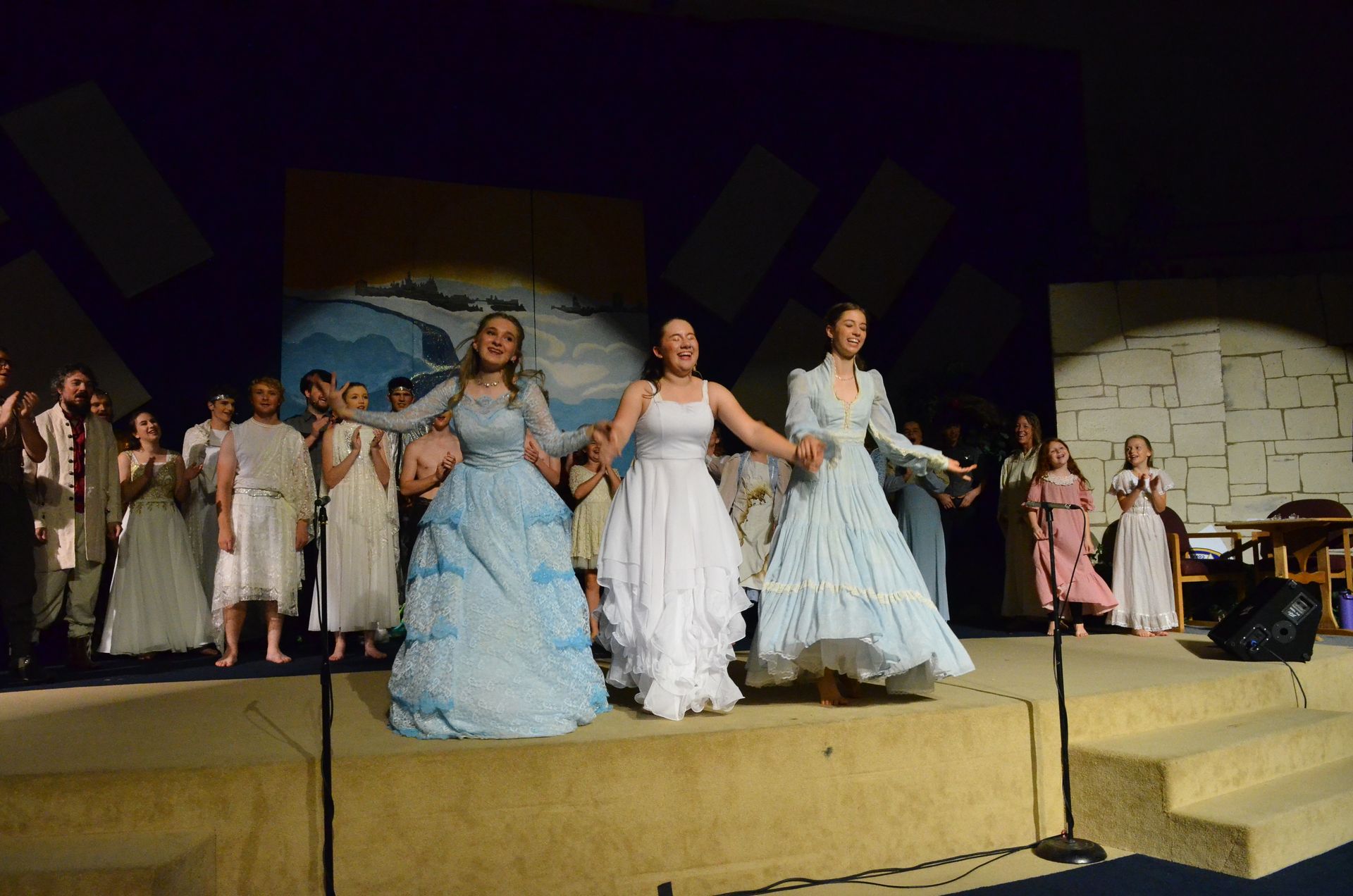 Three performers in flowing light blue and white gowns hold hands center stage while surrounded by cast members in period costumes, with painted backdrops and set pieces visible behind them on an illuminated theatrical stage.