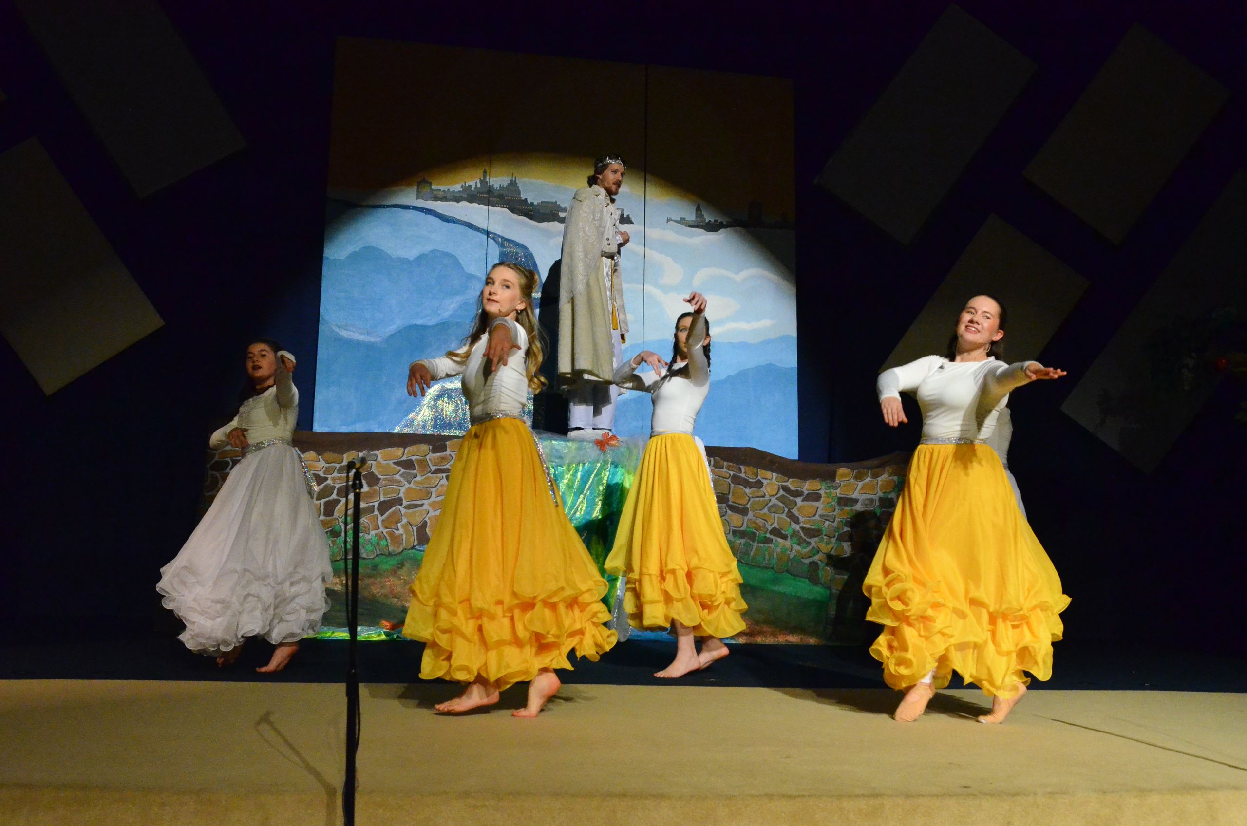 Four performers in white and yellow flowing skirts stand barefoot on stage before a painted backdrop depicting mountains and a distant castle, with the performers in various expressive poses.