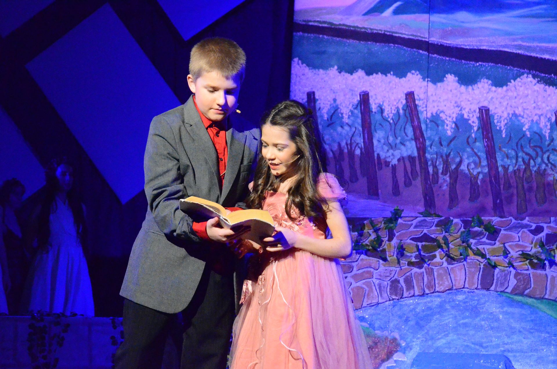 Two performers stand together on stage holding an open book, with the male performer in a gray blazer and red shirt and the female performer in a pink dress, set against a painted backdrop depicting a waterside landscape with wooden posts and blue-purple lighting.