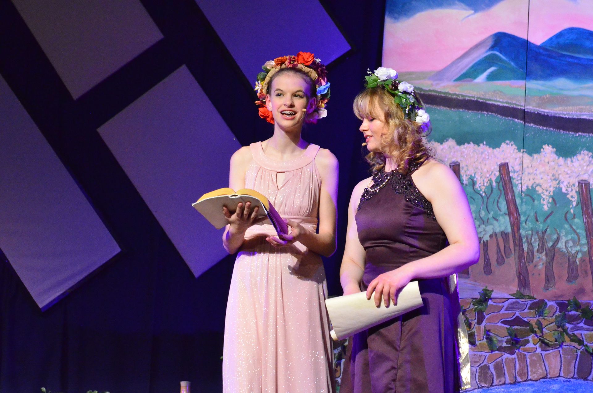 Two performers in flowing pastel gowns adorned with flower crowns stand center stage holding scripts, with stylized scenic projections of mountains and flowering trees displayed behind them.
