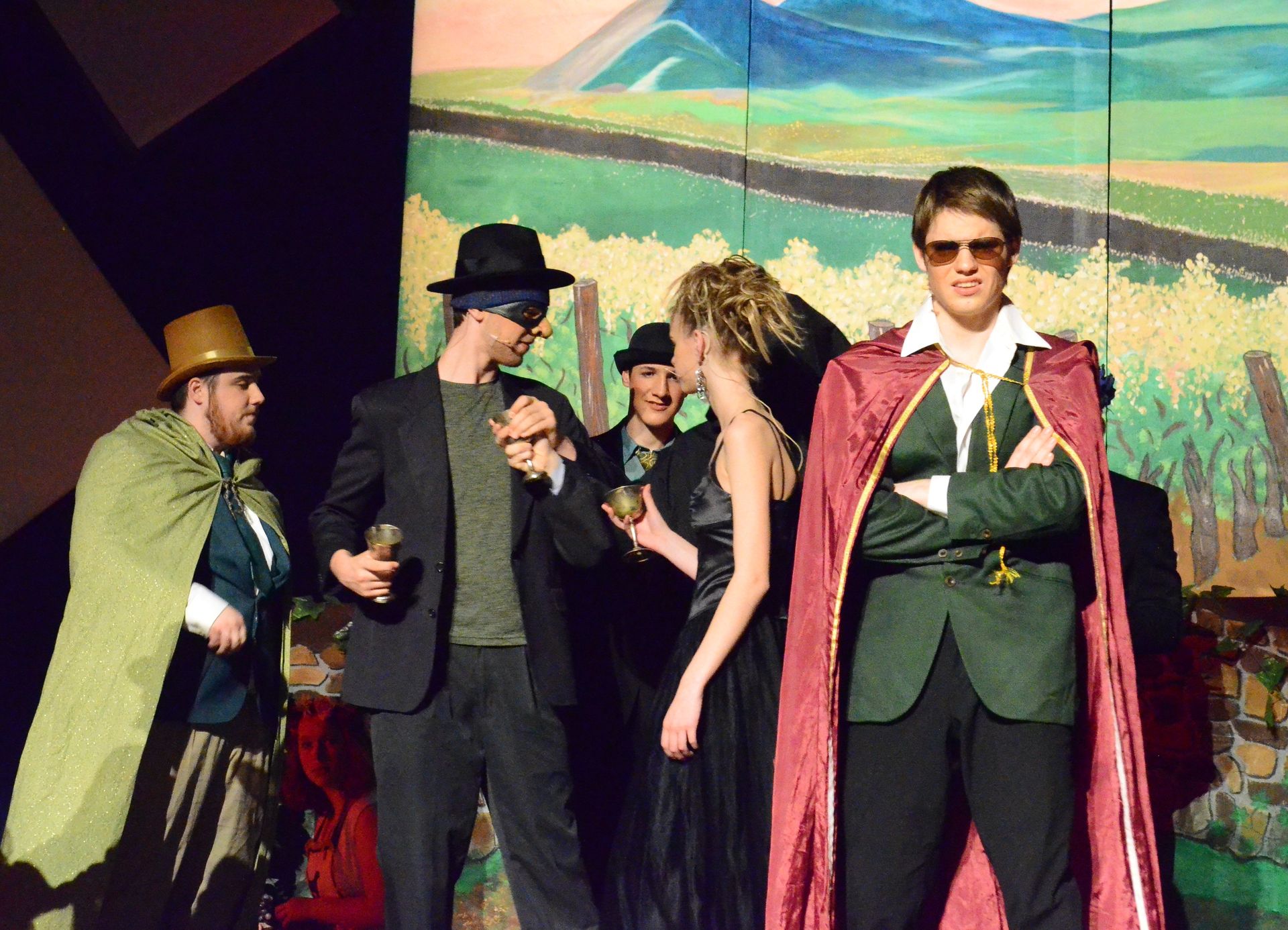 Six performers in colorful period costumes including capes, hats, and sunglasses stand together on stage in front of a painted backdrop depicting a pastoral landscape with fields and mountains.