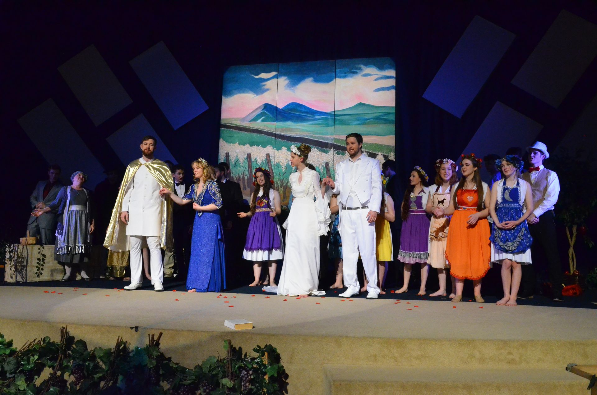 A diverse cast of performers in colorful period costumes stands on stage before a painted backdrop of mountains and sky, with geometric blue shapes suspended above them.