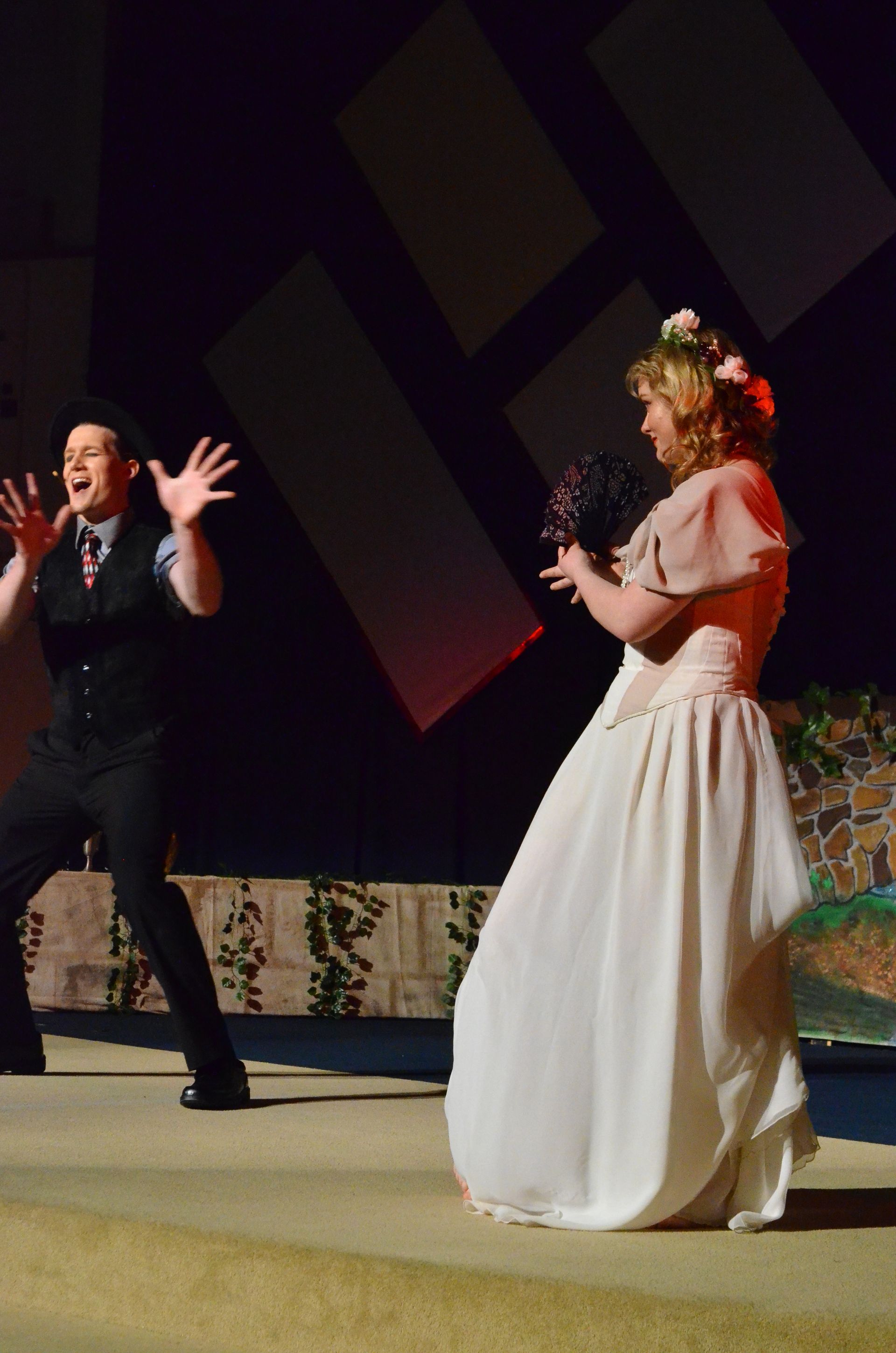 Two performers on stage, one wearing a white dress and floral headpiece and another in dark clothing, interact in a dramatic scene with stage lighting and decorative backdrop elements visible.