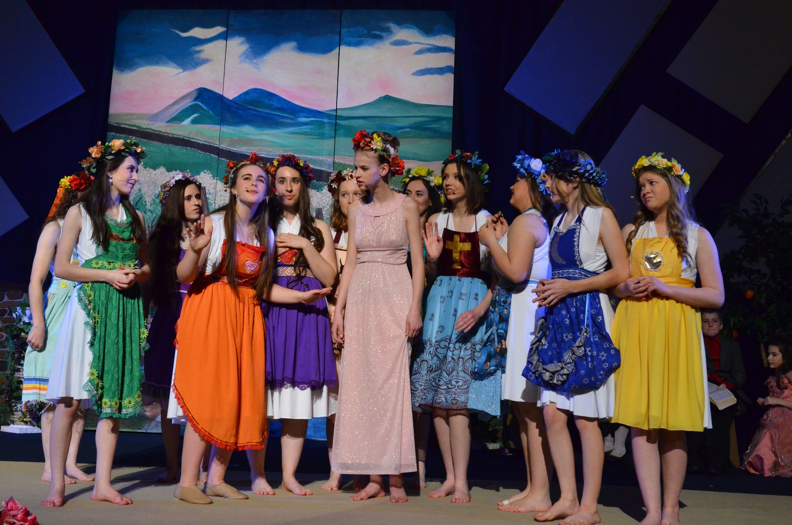 A cast of performers in colorful traditional dresses and flower crowns stands together on stage in front of a painted backdrop depicting mountains and sky.