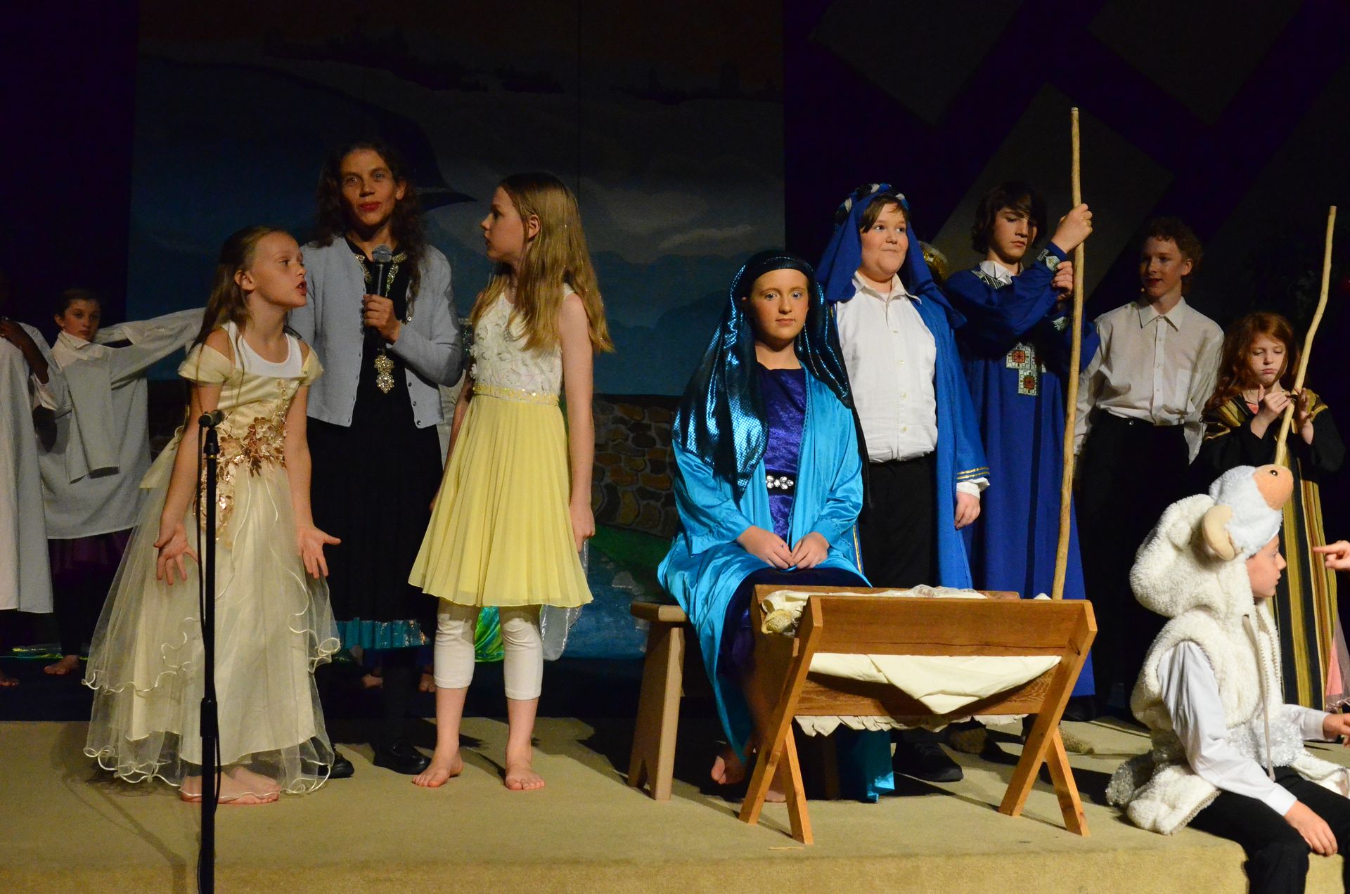 A cast of performers in period costumes stands on stage around a wooden manger, with some wearing blue robes and others in pale dresses, set against a painted backdrop of a night sky.