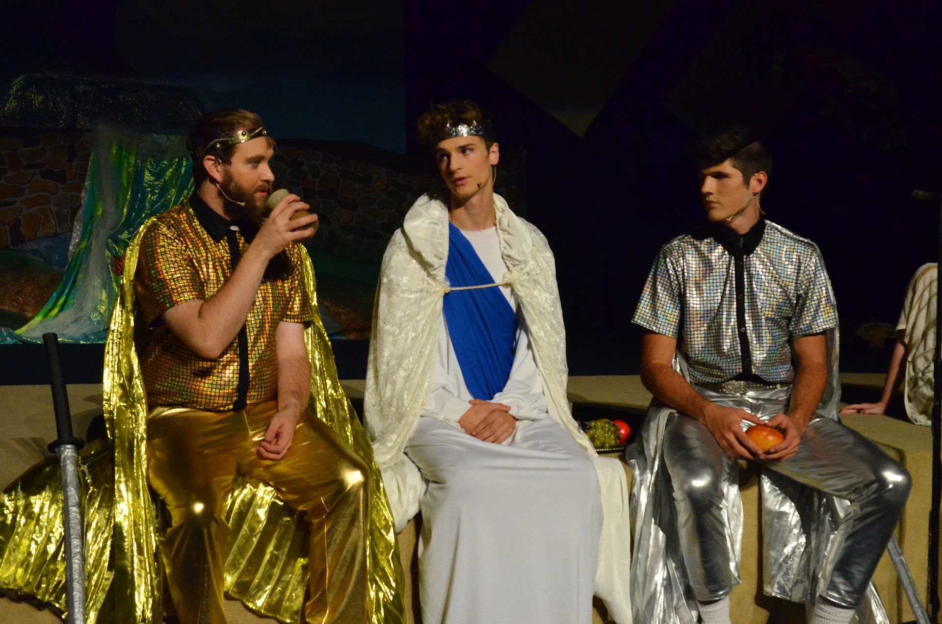 Three performers in gold, white, and silver metallic costumes sit on stage wearing laurel wreaths, with one holding a microphone, against a painted backdrop depicting an ancient Mediterranean setting.