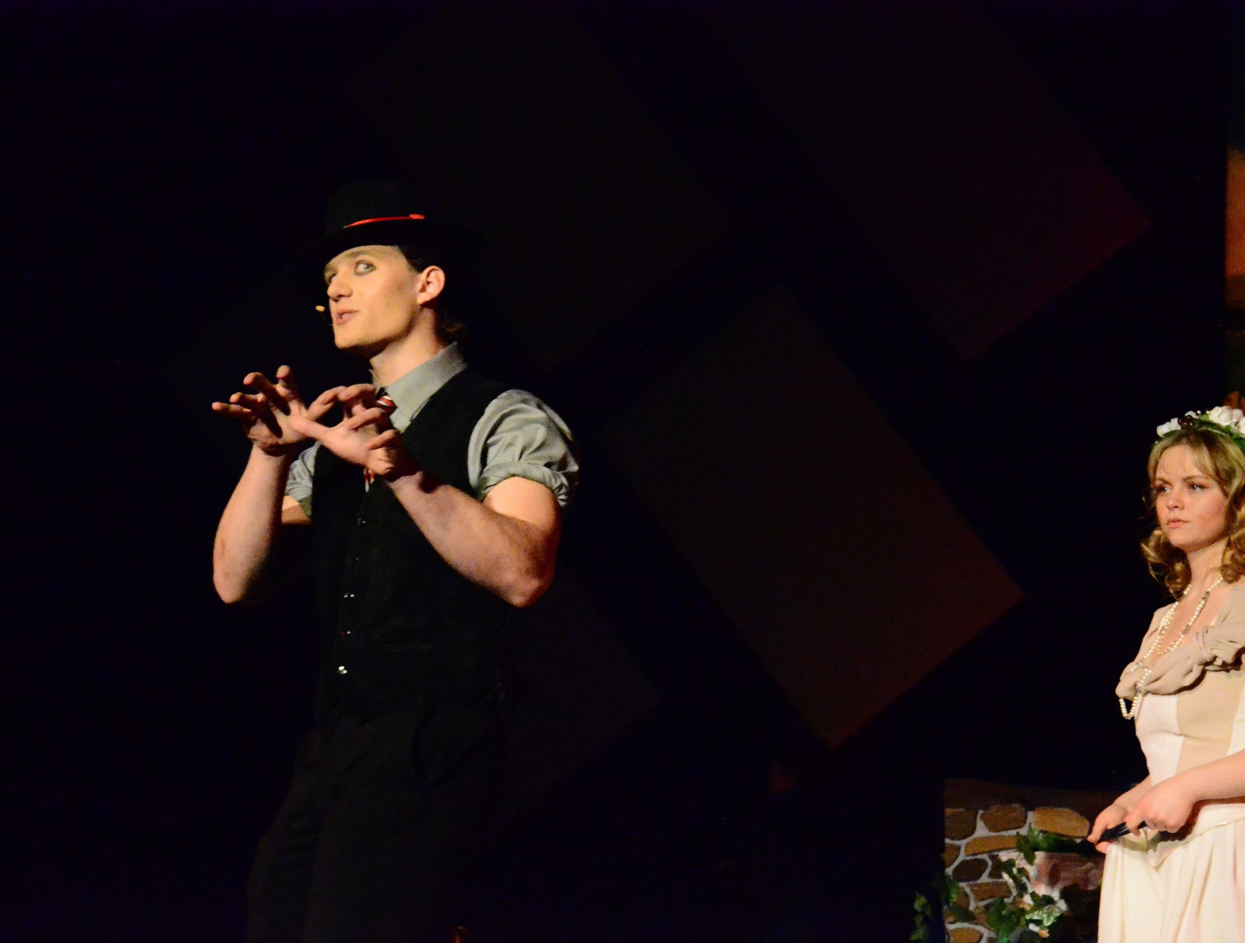 A performer in a black vest and cap gestures expressively while speaking on stage, with another performer in a cream-colored dress standing nearby against a dark background.