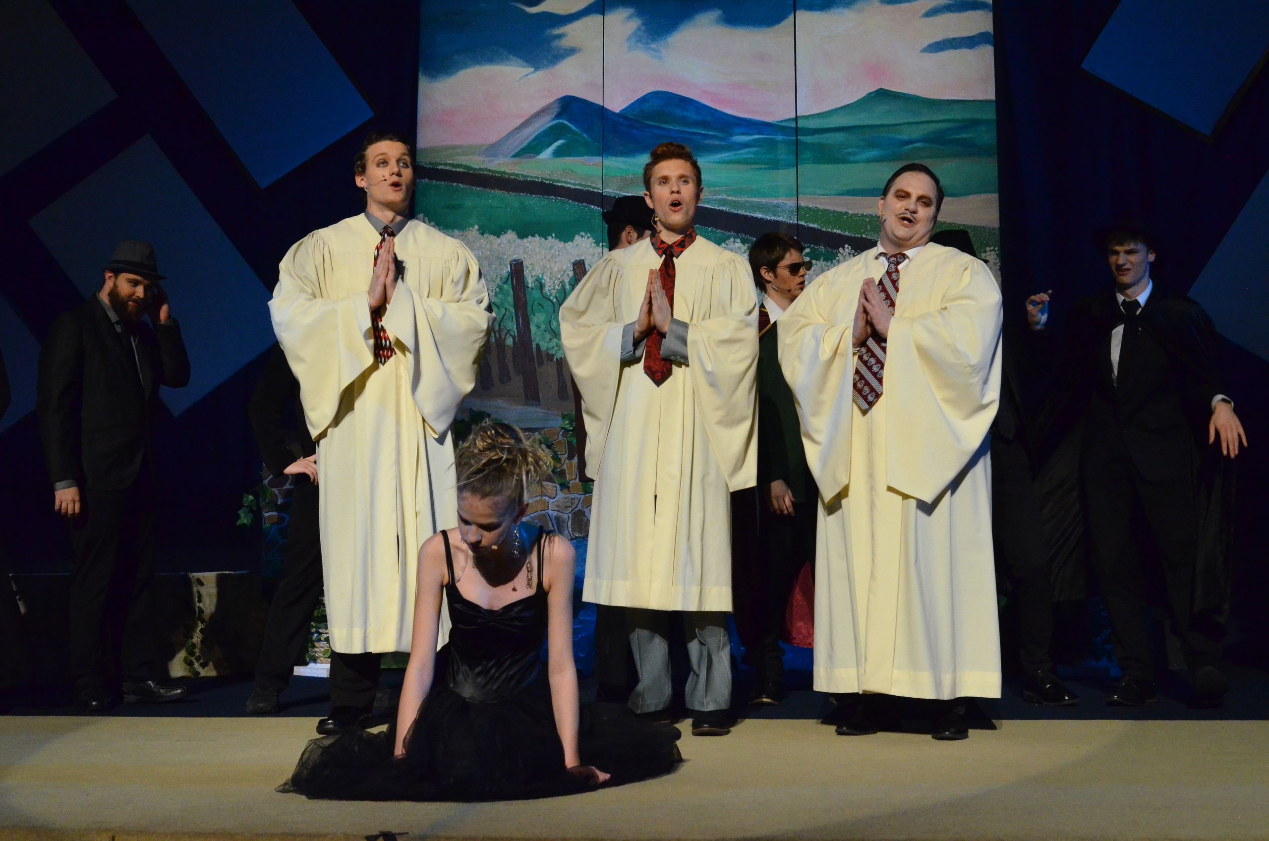 A cast of performers in cream-colored robes with colored ties stands on stage alongside others in dark formal attire, with one performer in a black dress kneeling in the foreground, all positioned before a painted backdrop depicting mountains and a pastoral landscape.