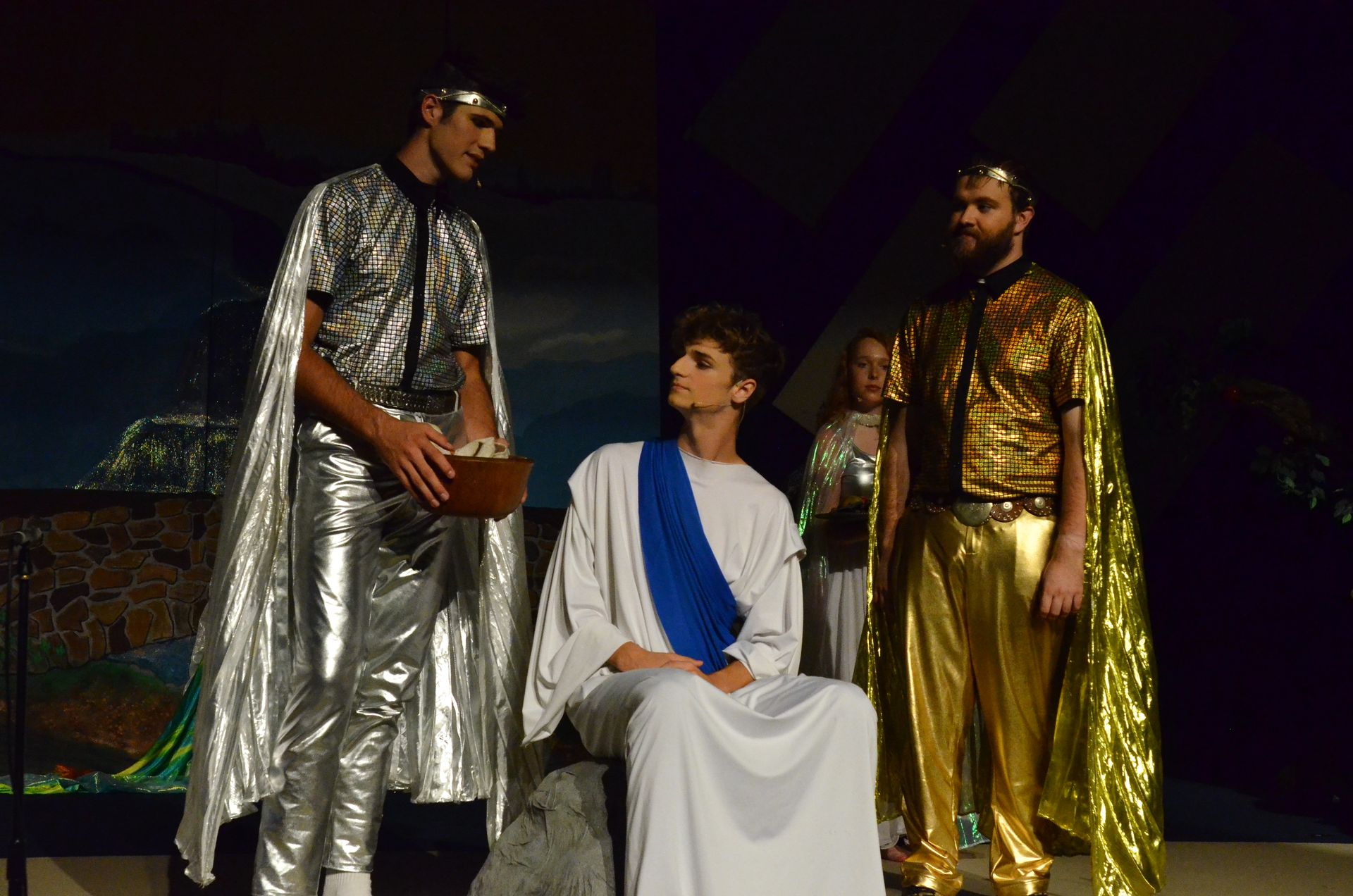 Four performers in elaborate metallic and flowing costumes stand on a darkened stage, with one seated in white robes and three standing around them in shimmering silver and gold garments, suggesting a scene of ancient or biblical significance.