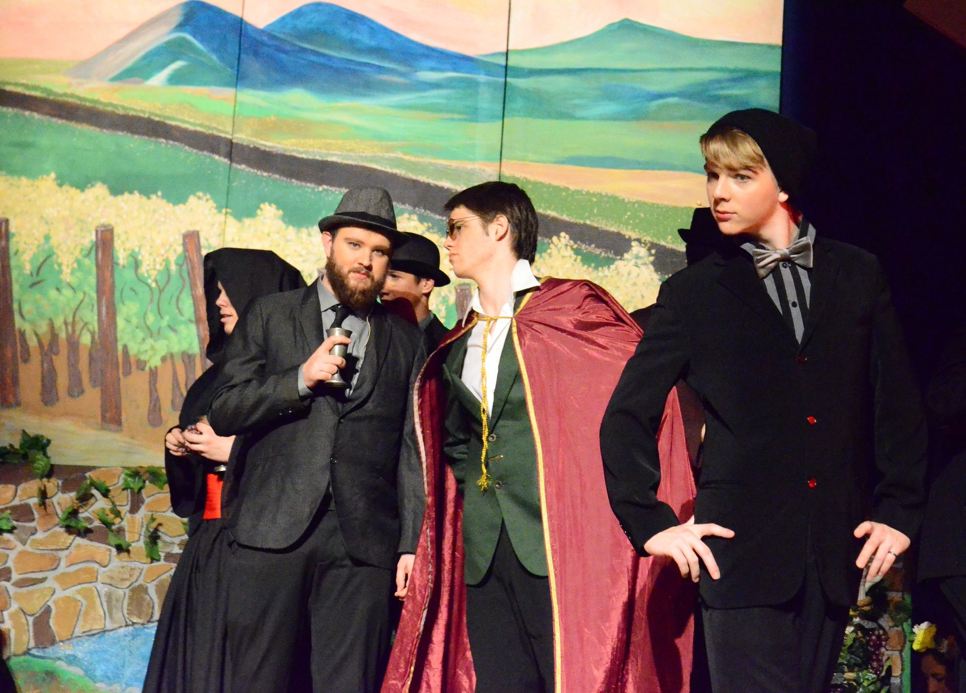 Five performers in period costumes stand on stage before a painted backdrop of mountains and fields, with one holding a microphone during what appears to be a scene from a theatrical production.