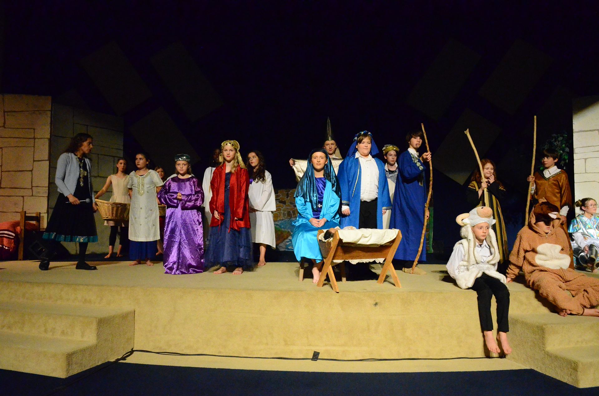 A cast of performers in biblical-era costumes stands on a tan stage set with wooden structures, with some seated figures positioned around a wooden manger in the center of the scene.