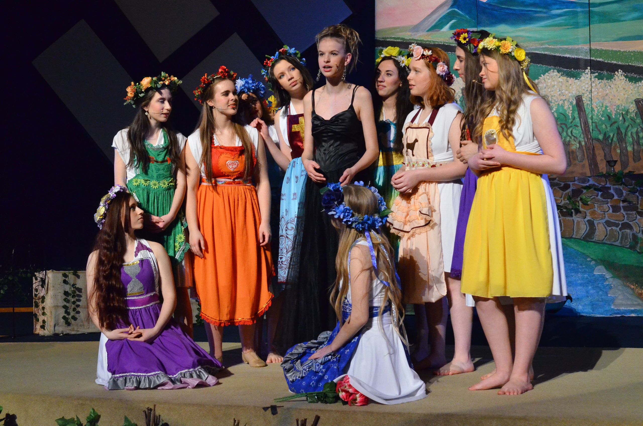 A cast of performers wearing colorful flower crowns and period costumes stands together on a stage with a painted backdrop of fields and sky, positioned as if in a dramatic scene.