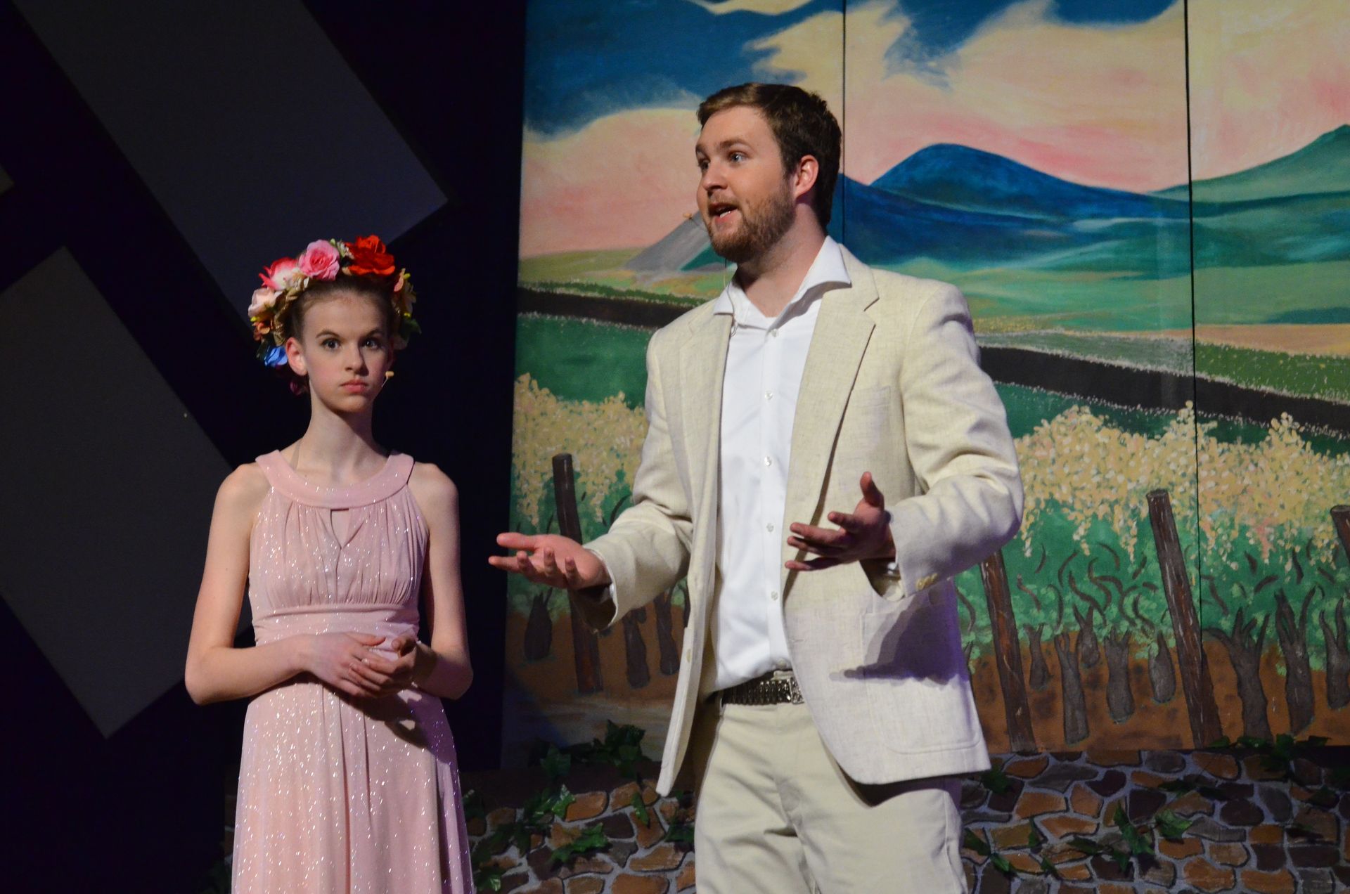 Two performers stand on stage in front of a painted landscape backdrop featuring rolling fields and mountains, with the performer on the left wearing a pink sleeveless dress with a floral crown and the performer on the right wearing a cream-colored blazer and white shirt while gesturing expressively.