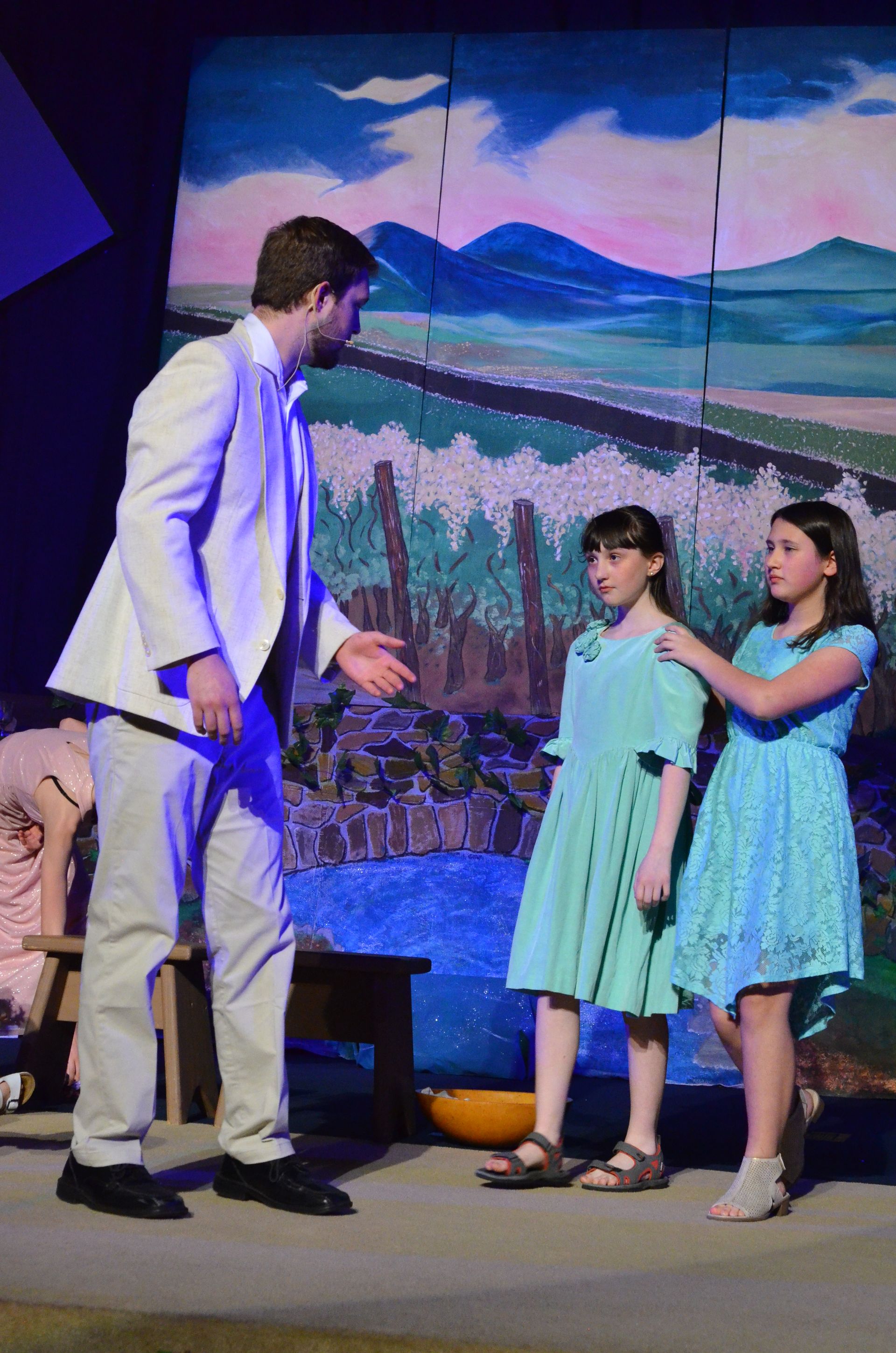 Four performers in blue and white costumes stand on stage before a vibrant projected backdrop of blues, greens, and earth tones depicting a natural landscape.