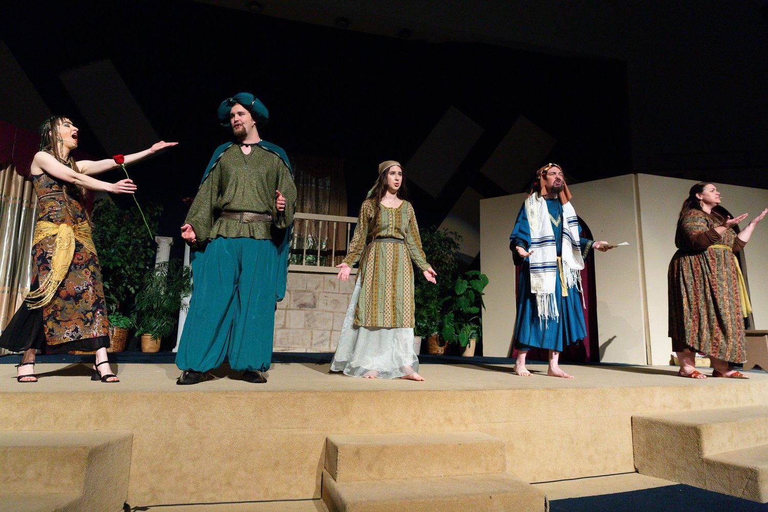 Five performers in period costumes stand on a staged platform with a stone building backdrop and potted plants, appearing to be performing in a biblical-themed musical production.