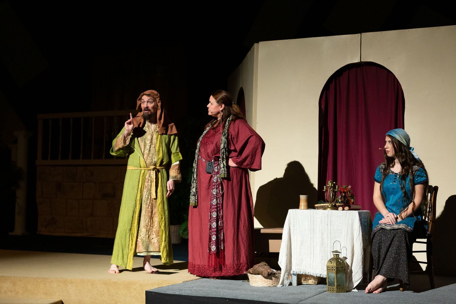Three performers in colorful period costumes perform a scene on stage, with one gesturing expressively while speaking, another standing nearby, and a third seated at a table with a white cloth, set against a backdrop featuring a burgundy arched doorway.
