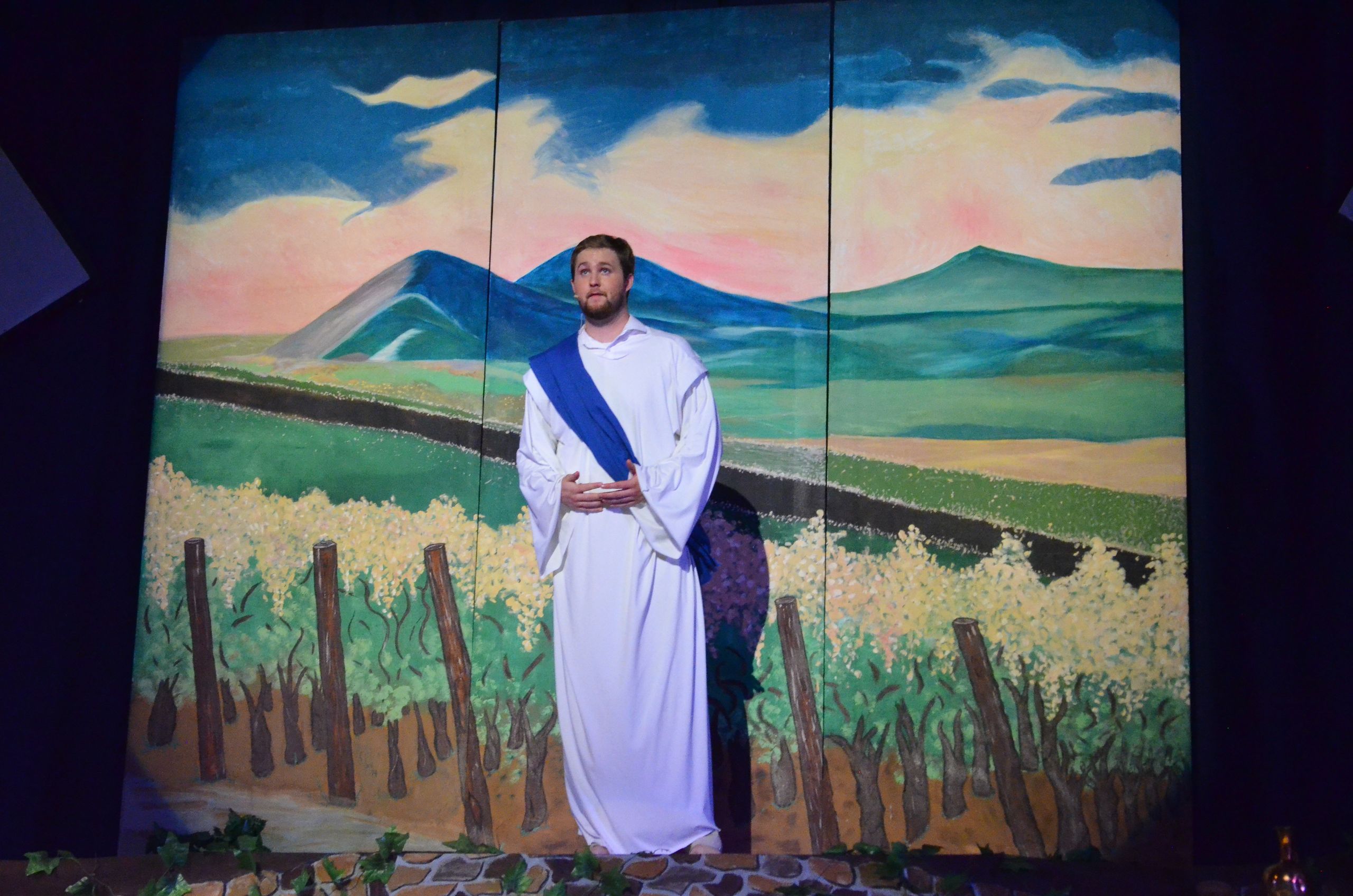A performer in a white robe and blue sash stands in front of a large painted backdrop depicting rolling mountains and a grain field with wooden fence posts, gazing thoughtfully into the distance.